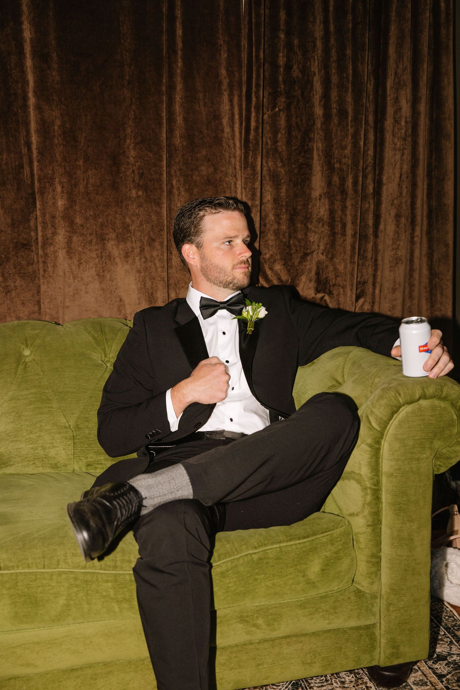 A groom in a black tuxedo sits on a green velvet sofa, holding a soda can, with a boutonniere on his lapel.