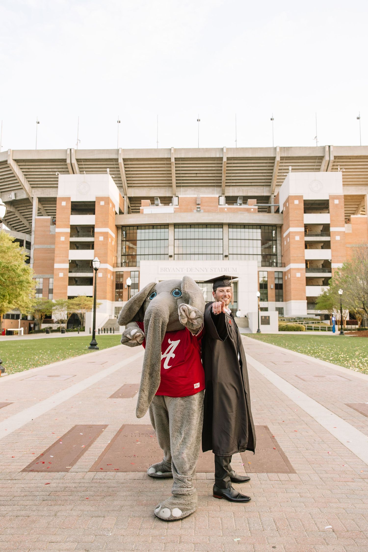 University of Alabama graduate poses with Big Al mascot statue in cap and gown outside Bryant-Denny Stadium.