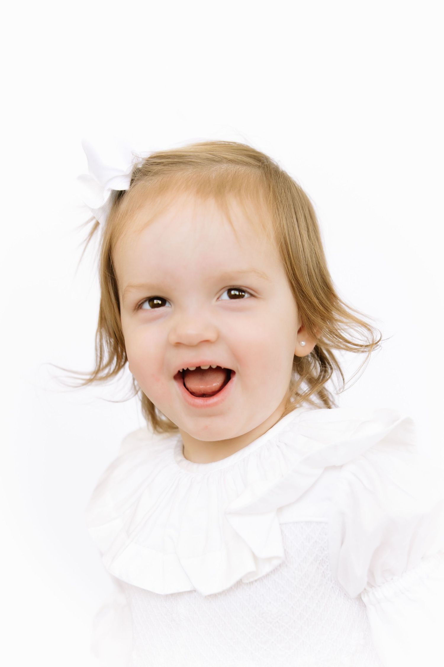 Adorable toddler girl laughing joyfully, wearing white dress and bow, portrait photo on white background.