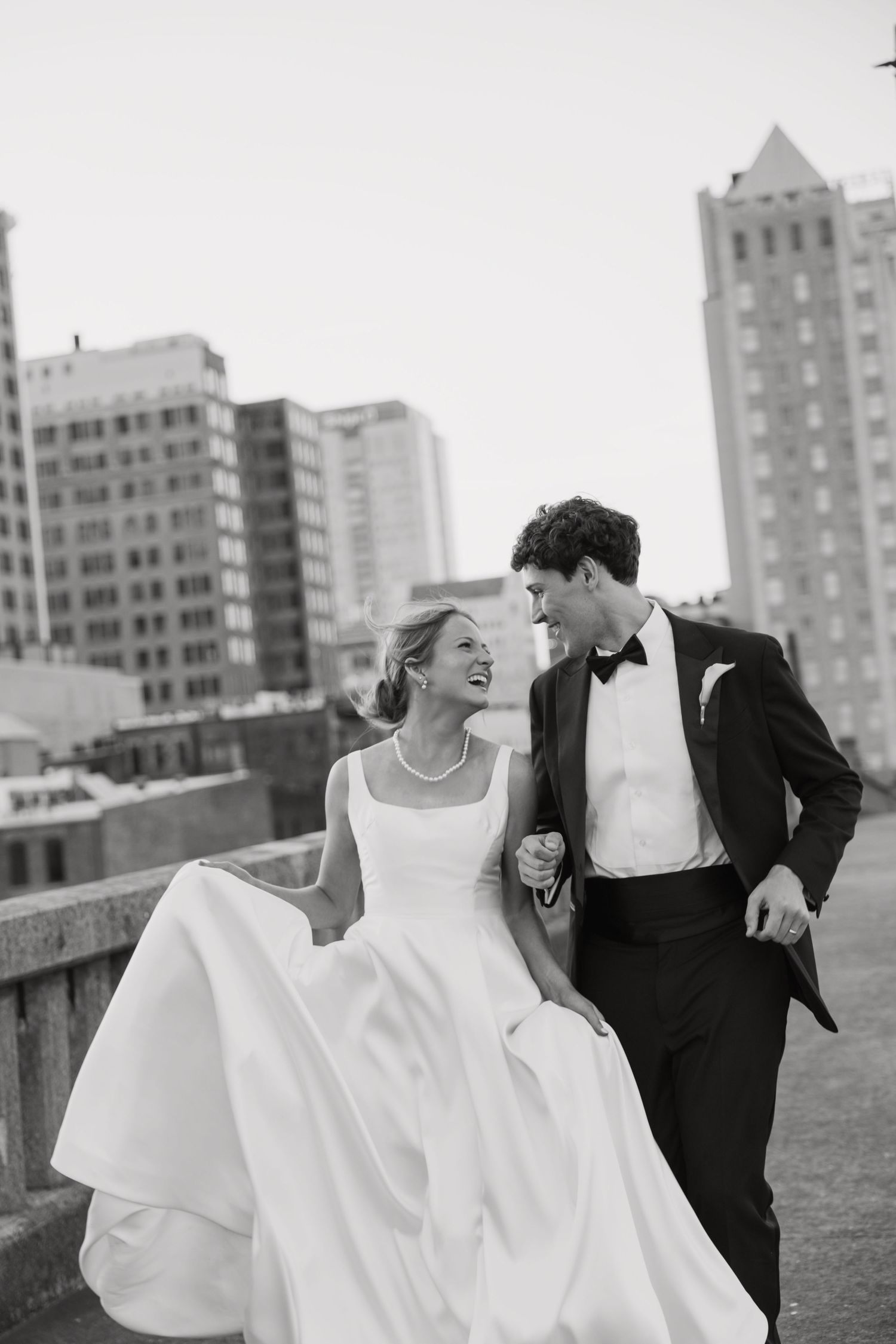 Black and white photo of laughing bride and groom walking arm in arm with city skyline behind them.