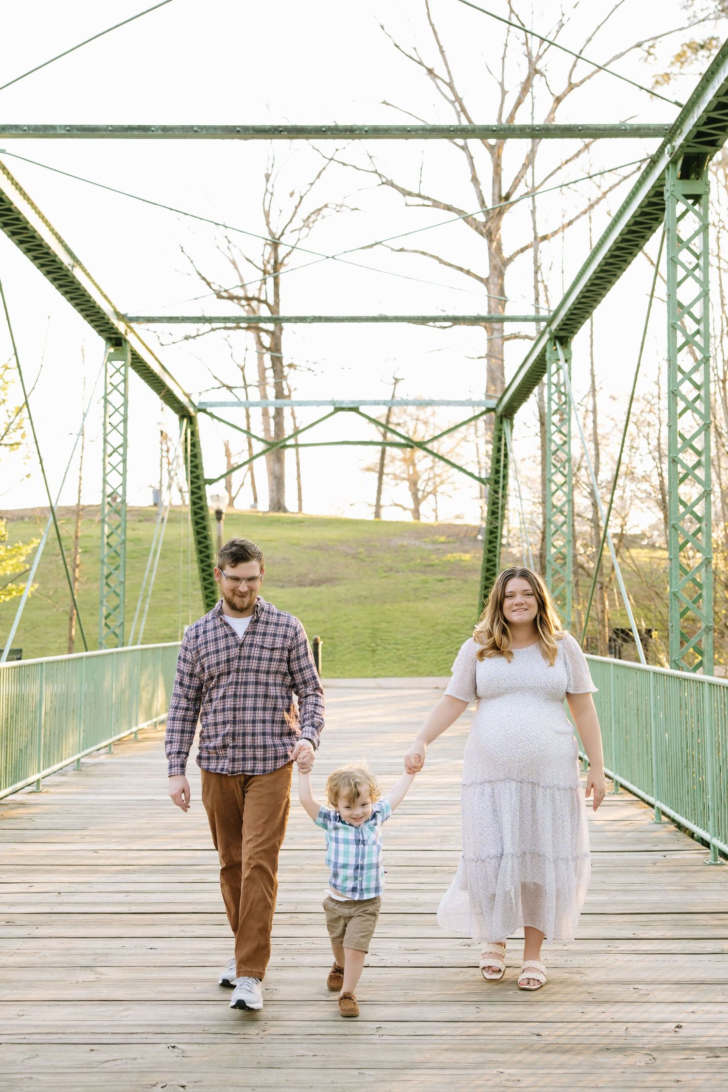 Pregnant woman in white dress walks with husband and toddler son on a green iron bridge outdoors.