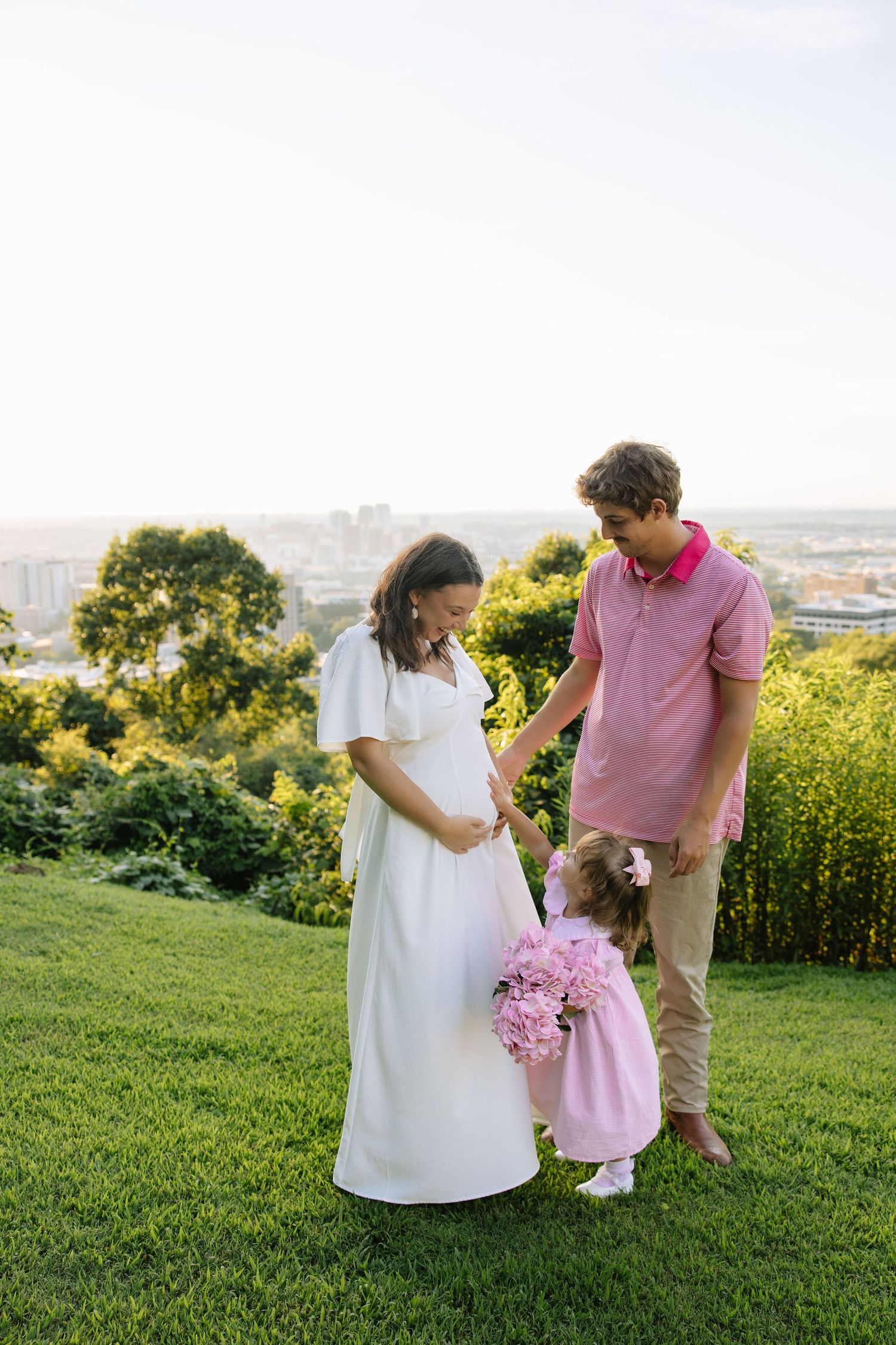 Pregnant woman in white dress with husband and daughter holding pink flowers on green hill at sunset.