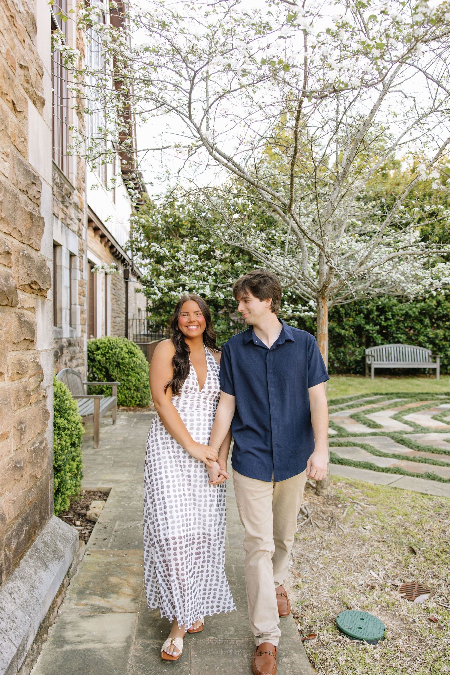 A couple walks hand-in-hand along a garden path beside a stone building, smiling at each other near a flowering tree.
