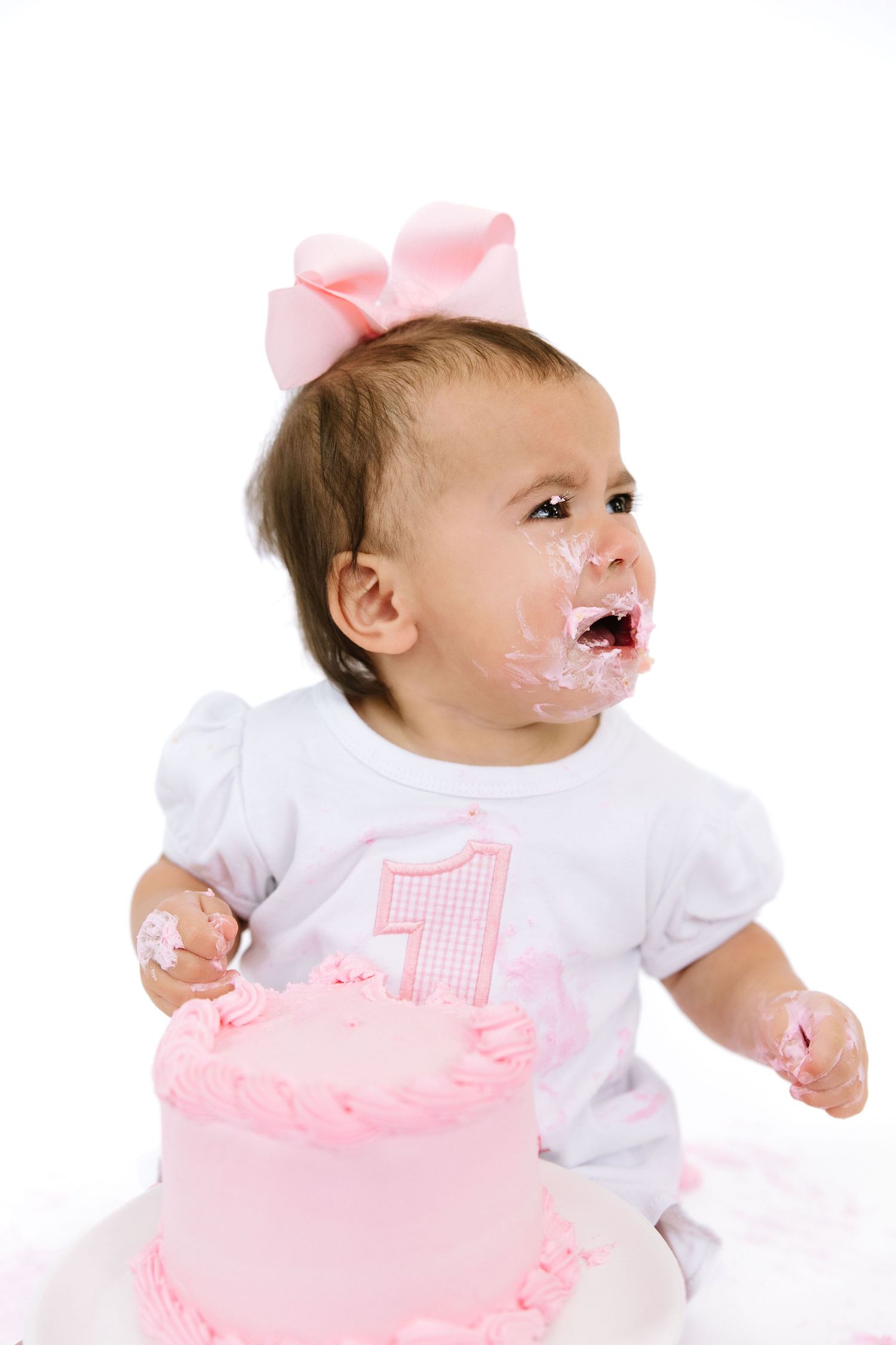 Baby girl with pink bow celebrates first birthday, face covered in frosting from pink smash cake.