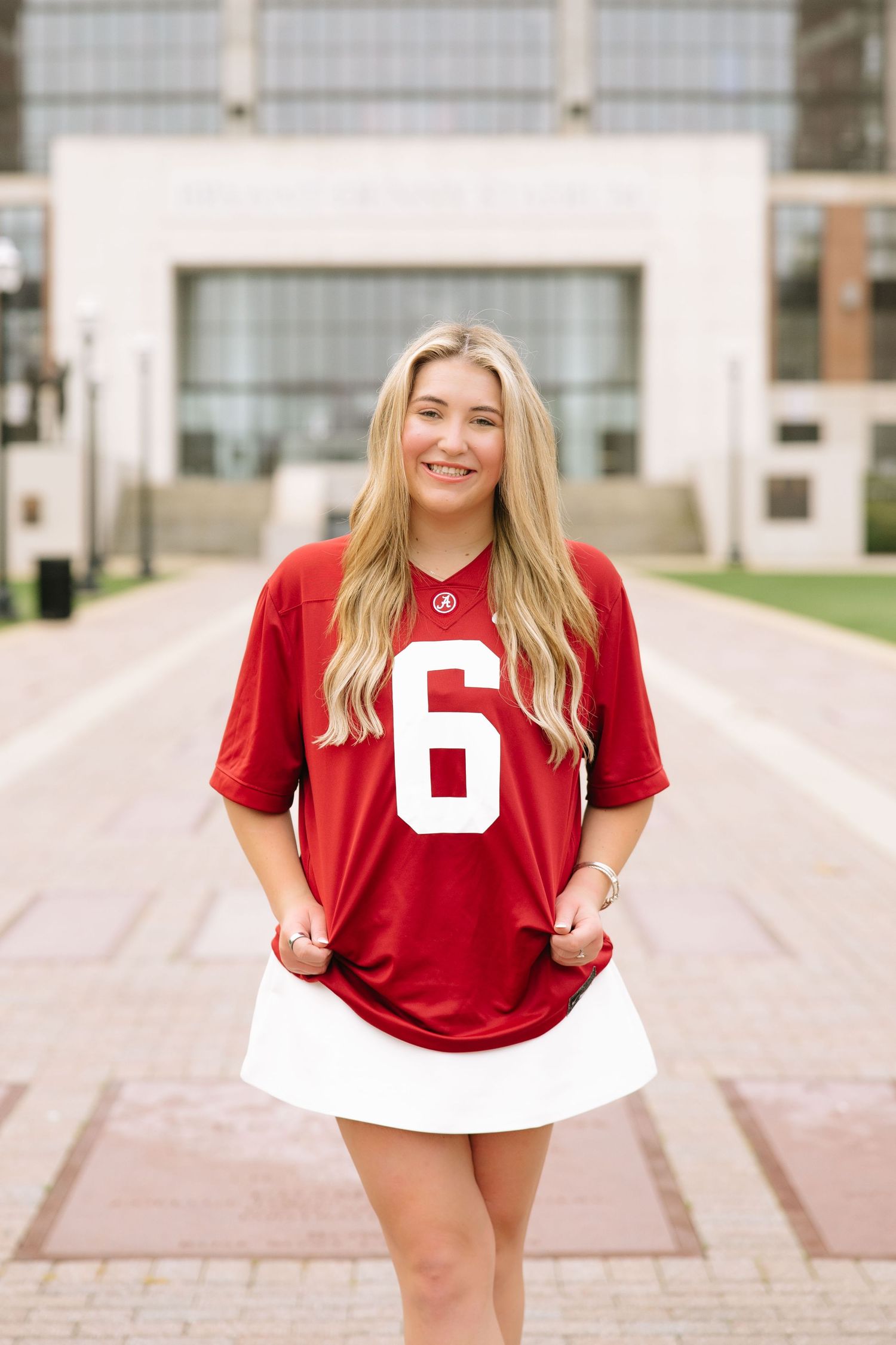 Smiling blonde woman wearing a red Alabama football jersey number 6 and white skirt on a university campus.