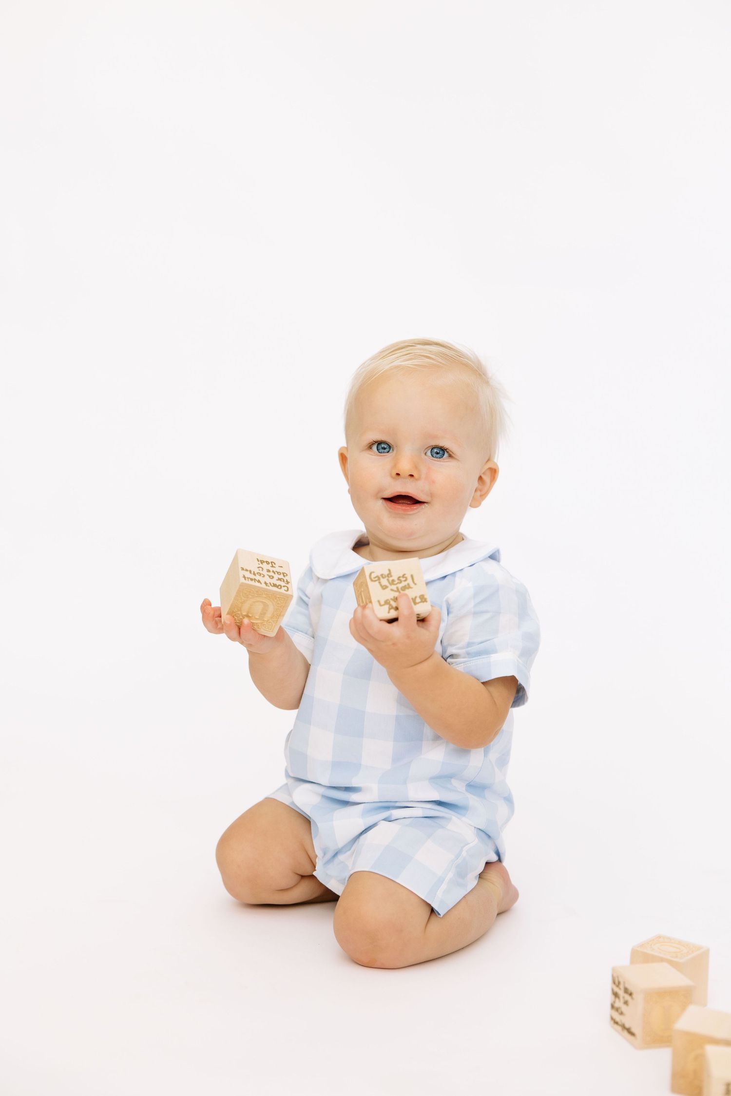Blonde baby with blue eyes in plaid romper sitting on white background playing with wooden alphabet blocks.