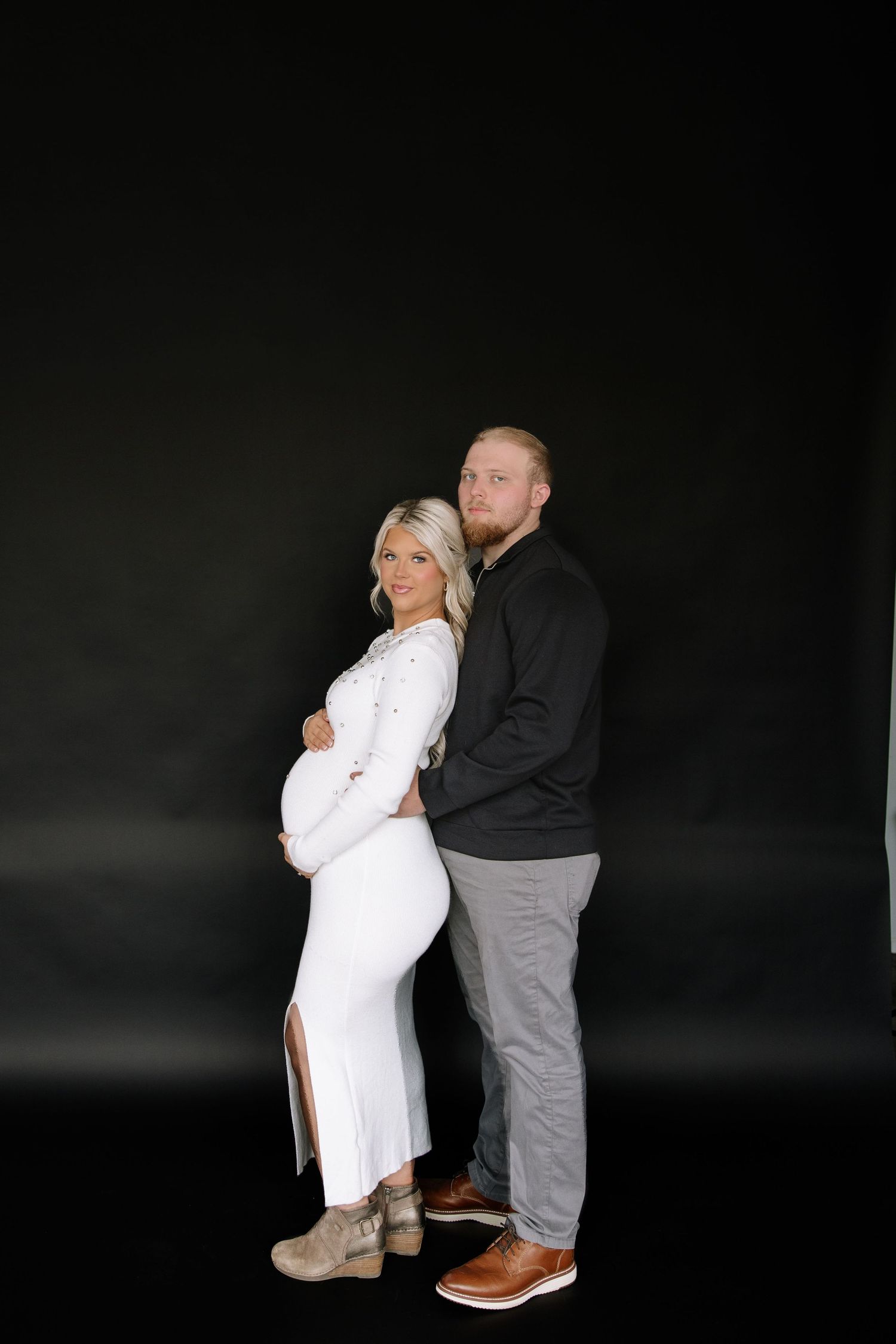 Pregnant woman in white dress poses back-to-back with bearded man in black shirt against dark studio background.