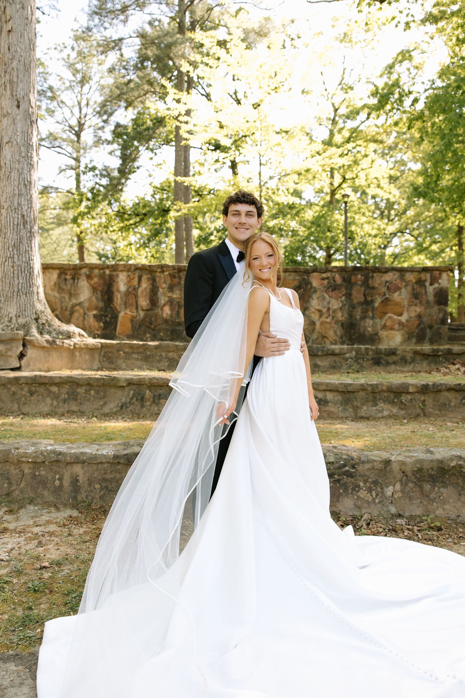 Bride and groom pose outdoors in wedding attire, her white gown and veil trailing gracefully amid lush green trees.