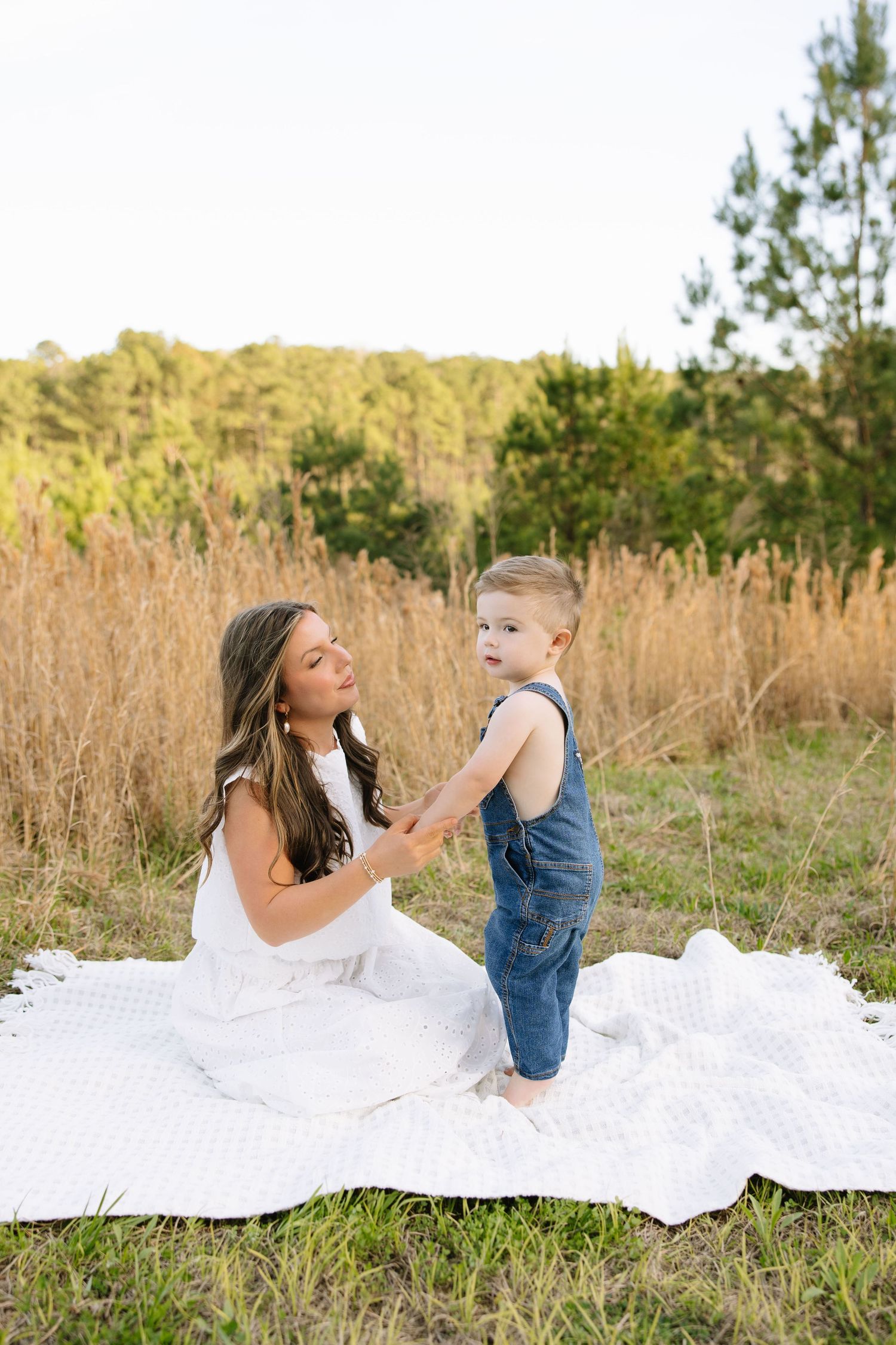 A mother in a white dress sits on a blanket outdoors with her toddler son in denim overalls.