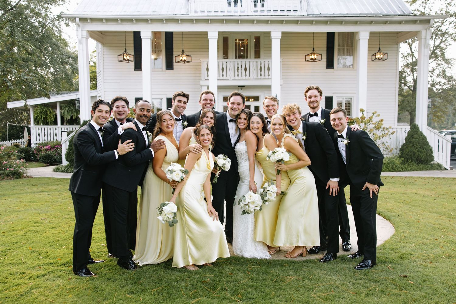 Wedding party in black tuxedos and pale yellow dresses pose in front of white southern mansion.