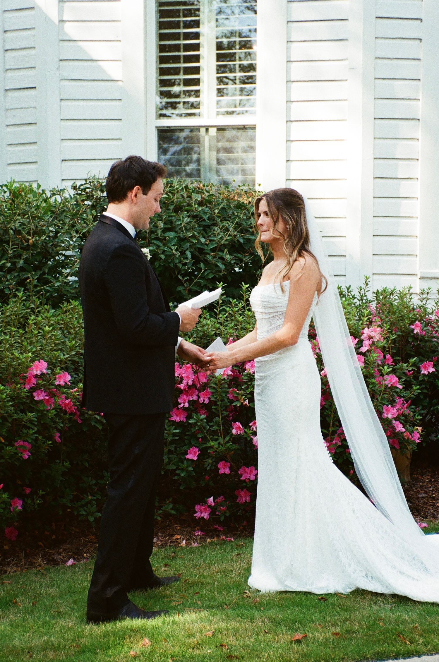 A bride and groom playfully dance and twirl in a garden with pink flowers and white shutters in the background.