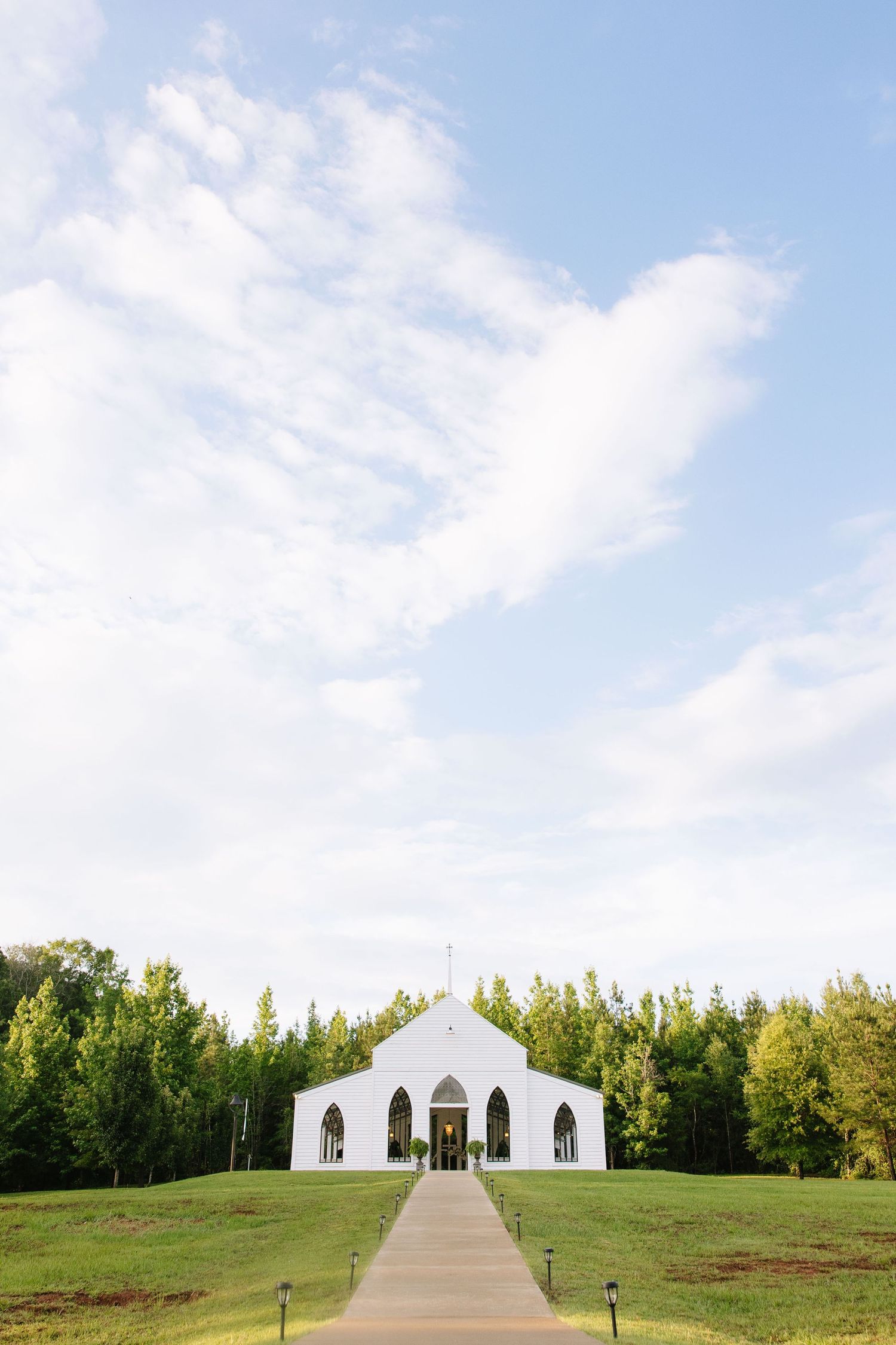 White chapel with arched windows and a paved pathway surrounded by lush green trees under a partly cloudy sky.