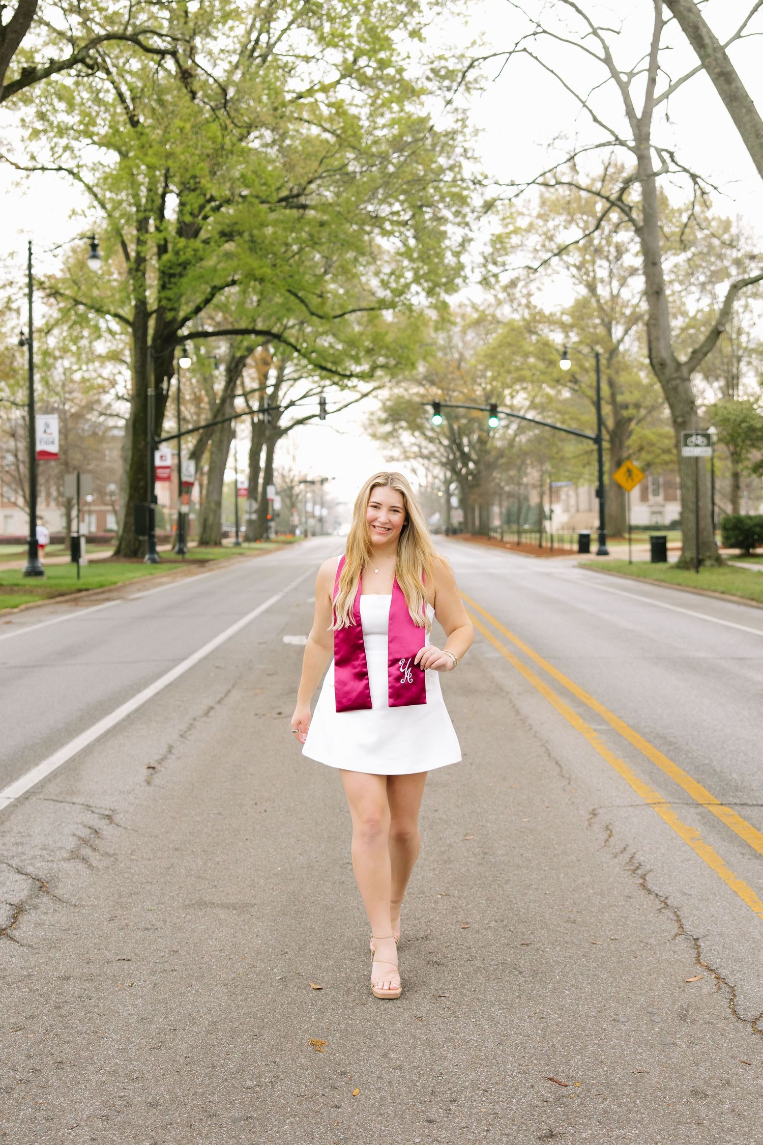 Smiling blonde woman in white dress holding pink clutch, posing on tree-lined street.