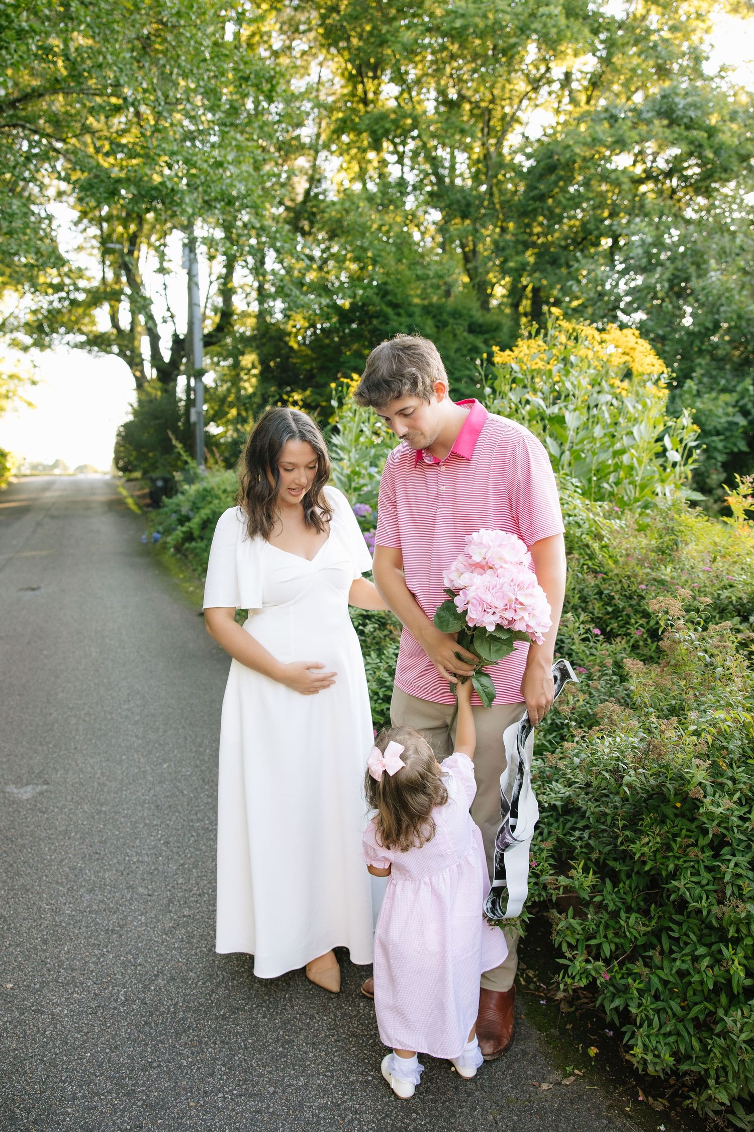 Pregnant woman in white dress with husband in pink polo and toddler daughter holding pink hydrangeas outdoors.