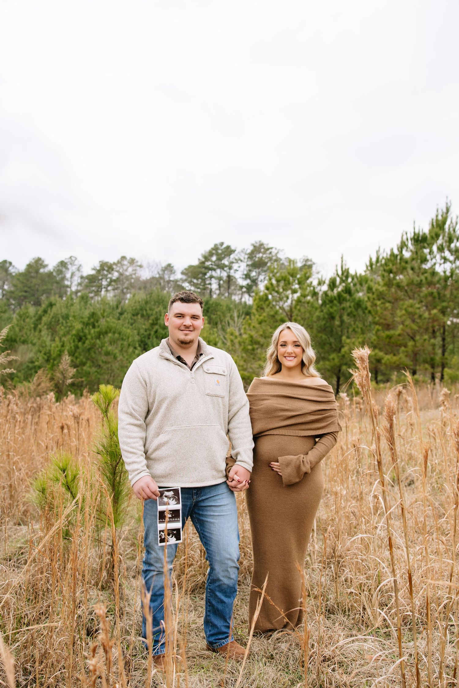Expecting couple holding ultrasound photos in golden field, woman in olive wrap maternity dress, man in cream pullover and jeans.