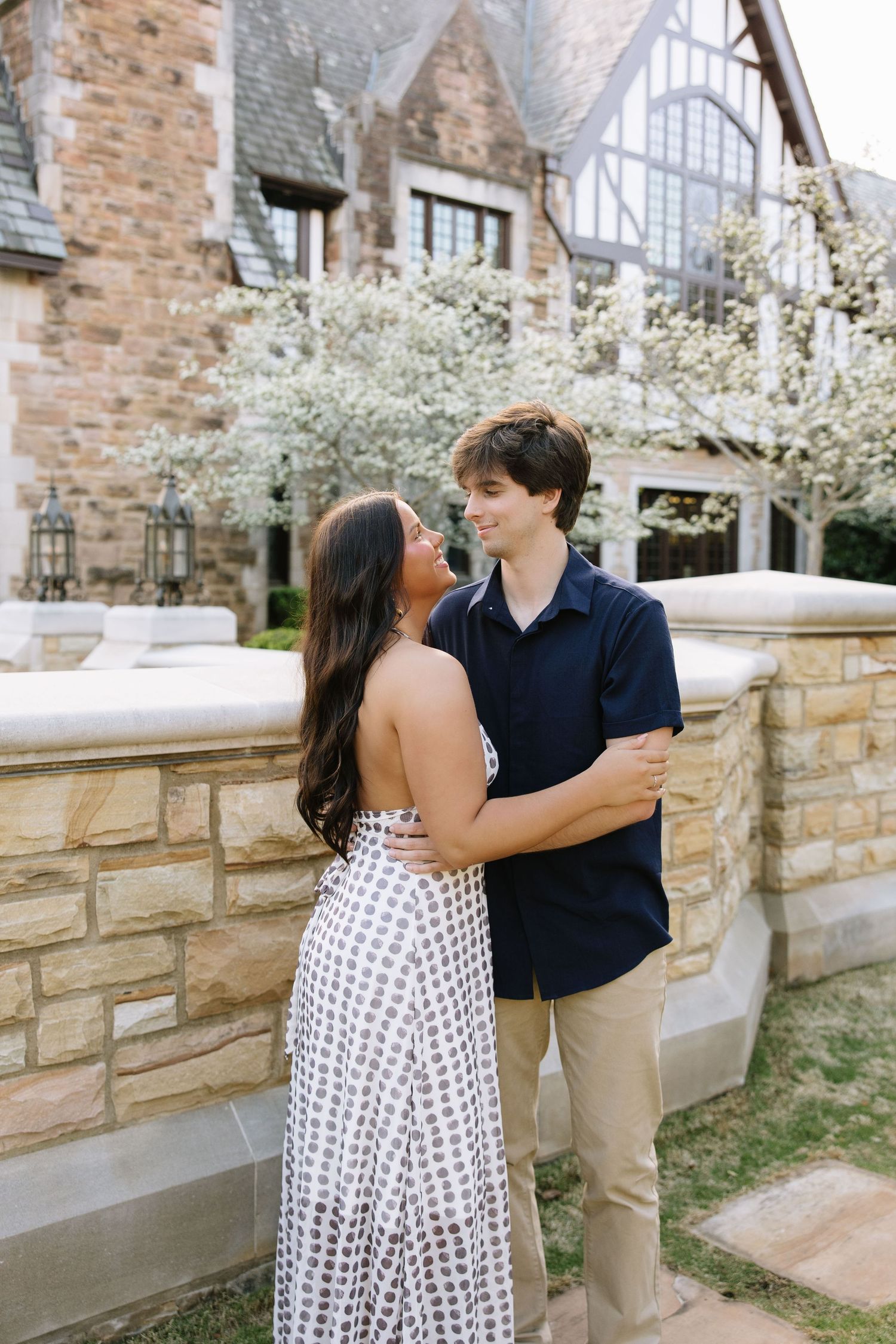 A young couple embraces outdoors near a stone wall with blooming white trees and a Tudor-style building behind them.