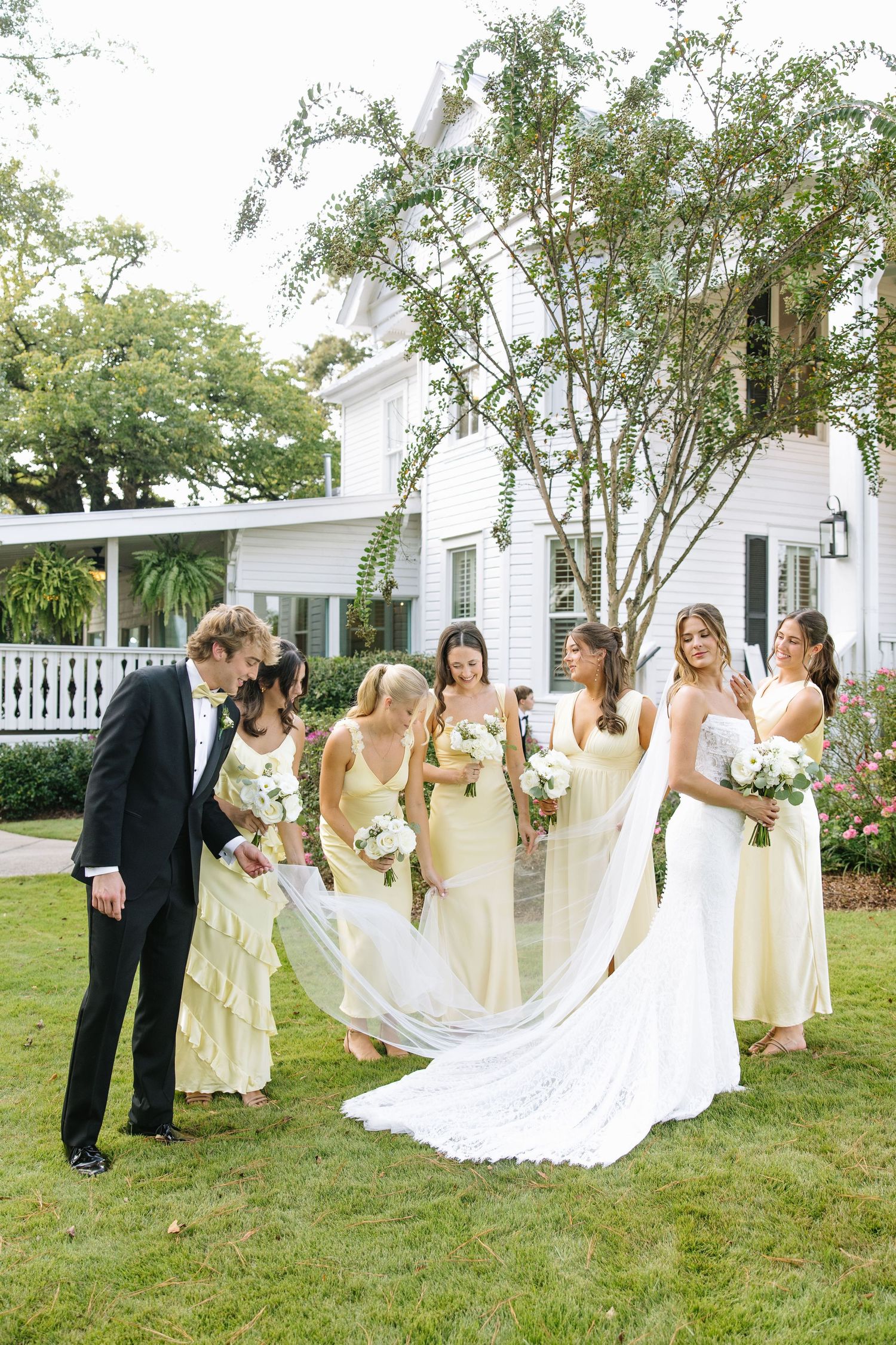 Wedding party in yellow dresses helps adjust a flowing white bridal gown train on a grassy lawn.