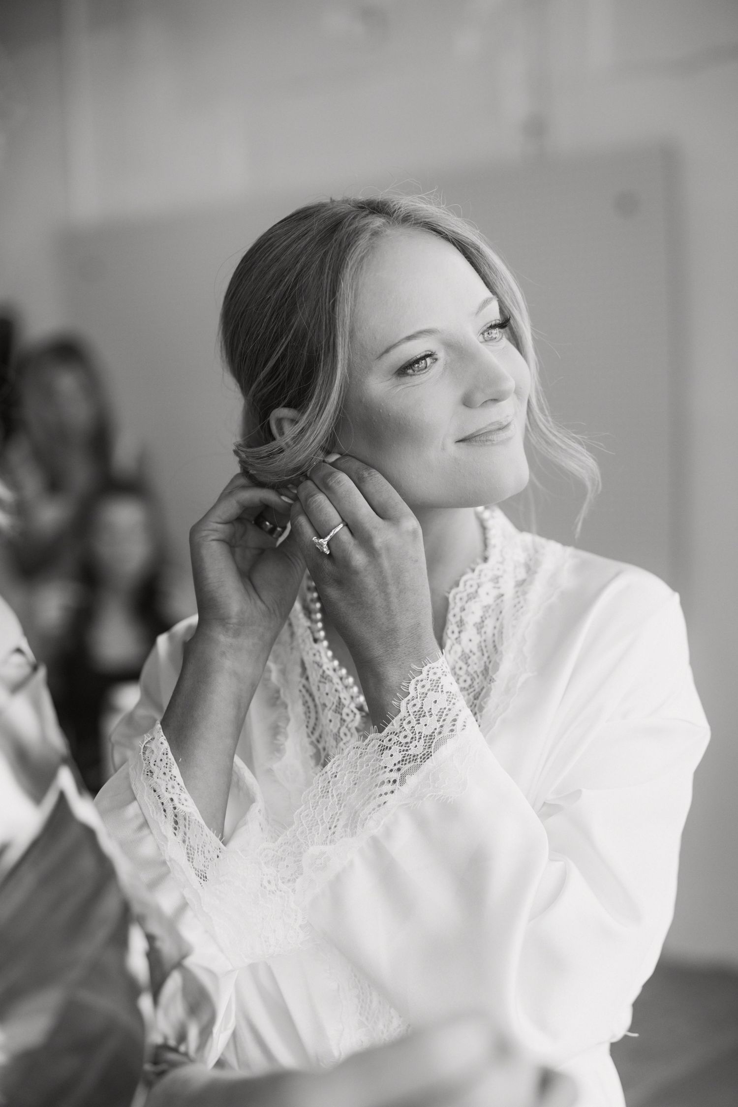 Black and white photo of a smiling bride putting on earrings while wearing a white lace robe.