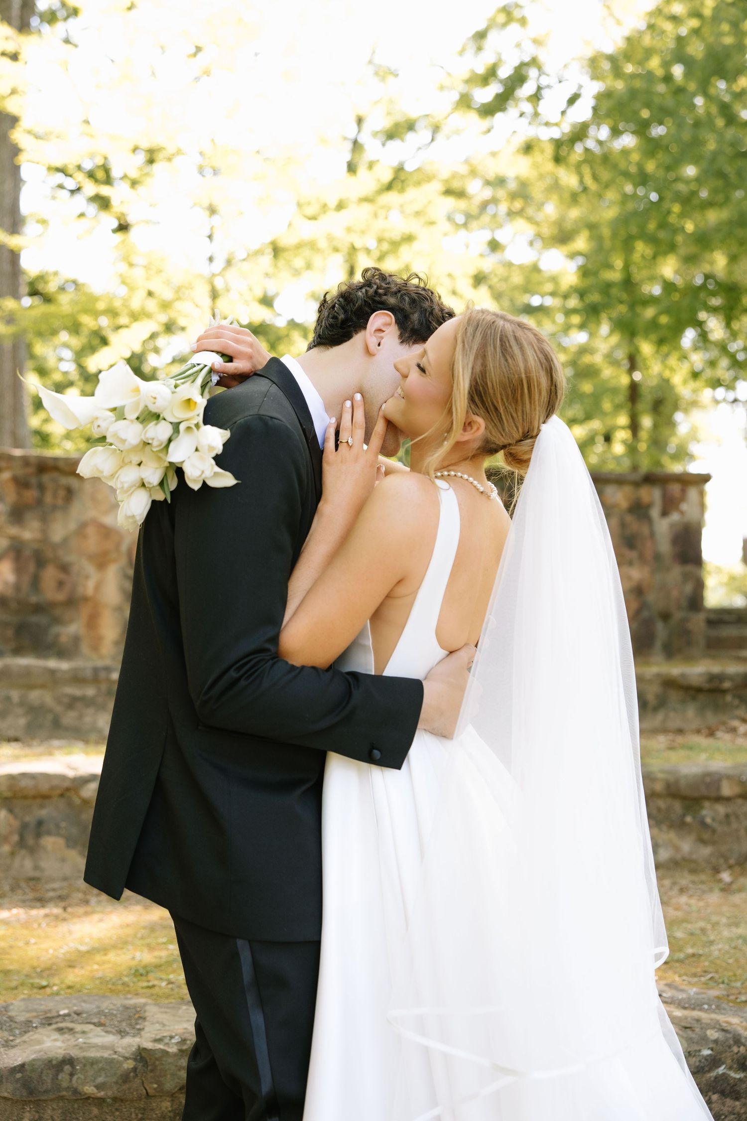 Bride and groom sharing a romantic kiss outdoors, bride holding white lily bouquet in elegant wedding attire.