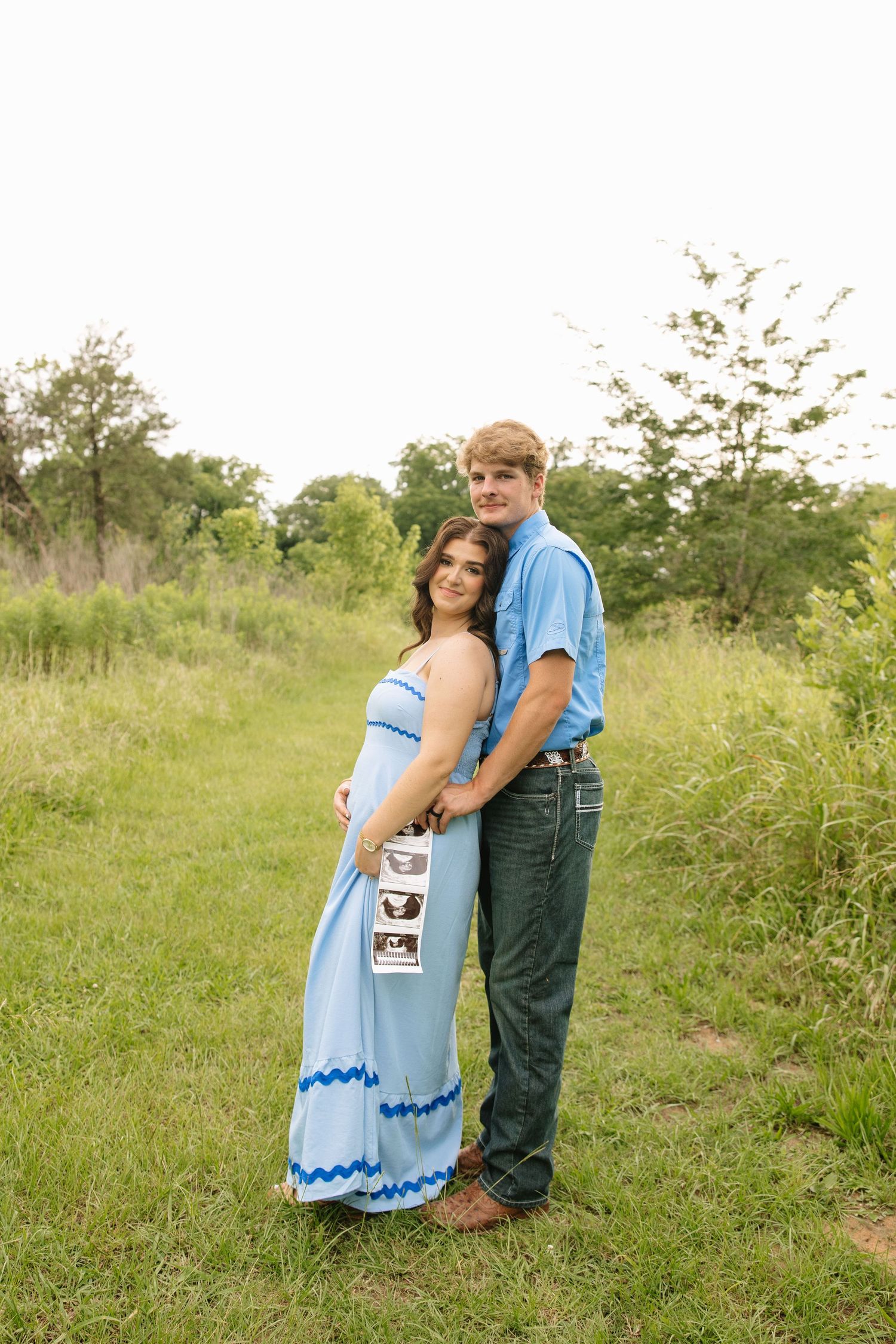 A couple poses outdoors in matching blue outfits, holding ultrasound photos, announcing their pregnancy in a green field.