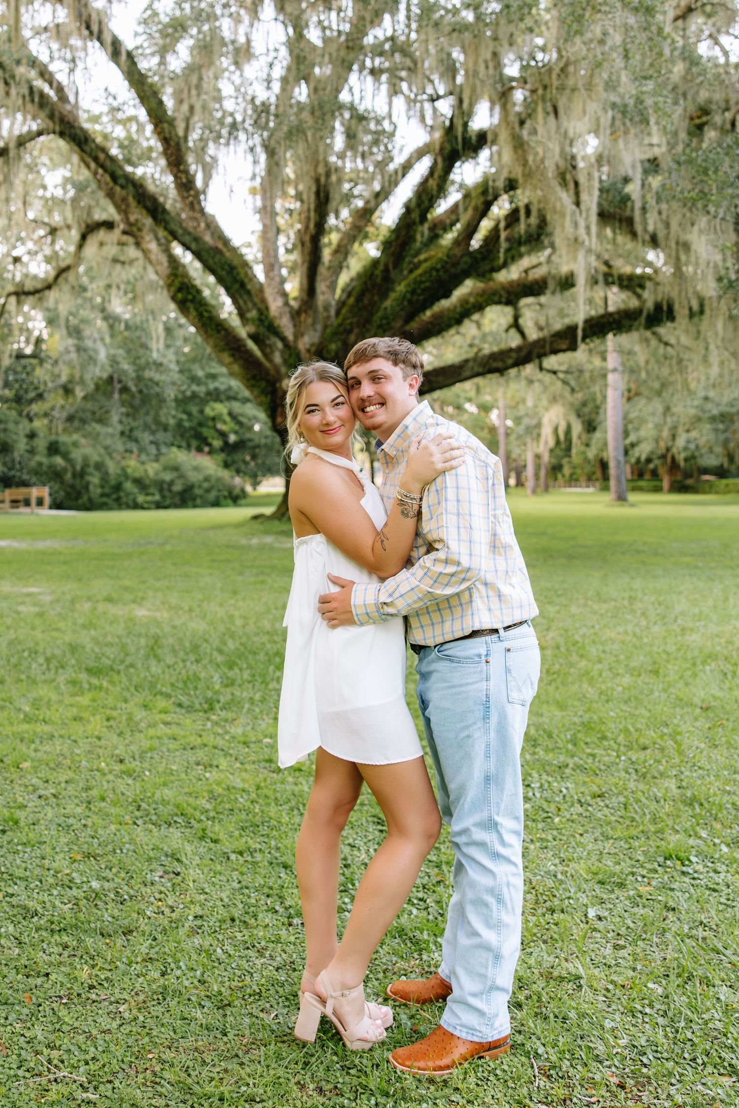 Couple embracing outdoors under a large Spanish moss-covered oak tree on green grass, woman in white dress, man in plaid shirt.
