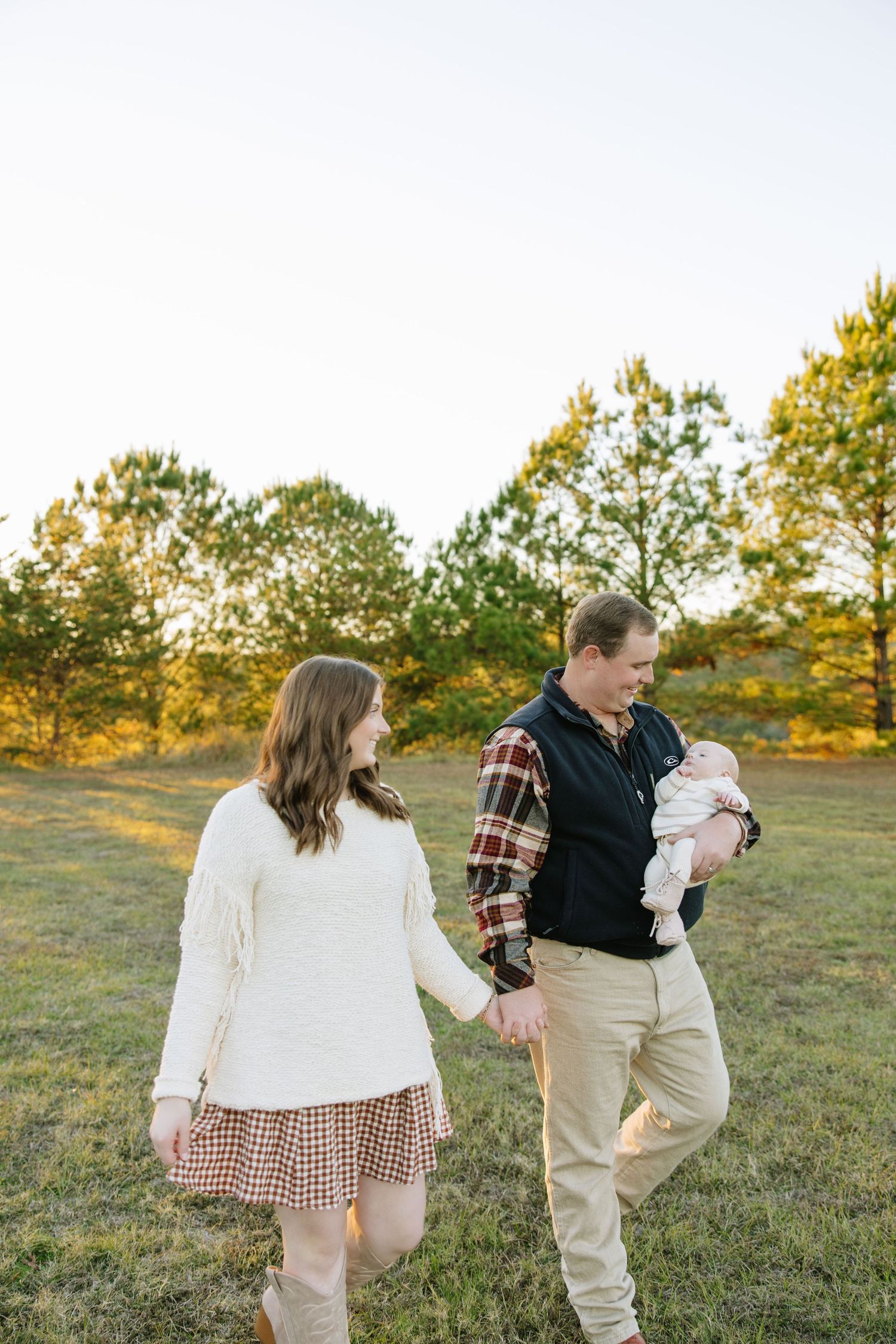 A couple walks hand-in-hand in a sunlit field, the man cradling a newborn baby, surrounded by autumn trees.