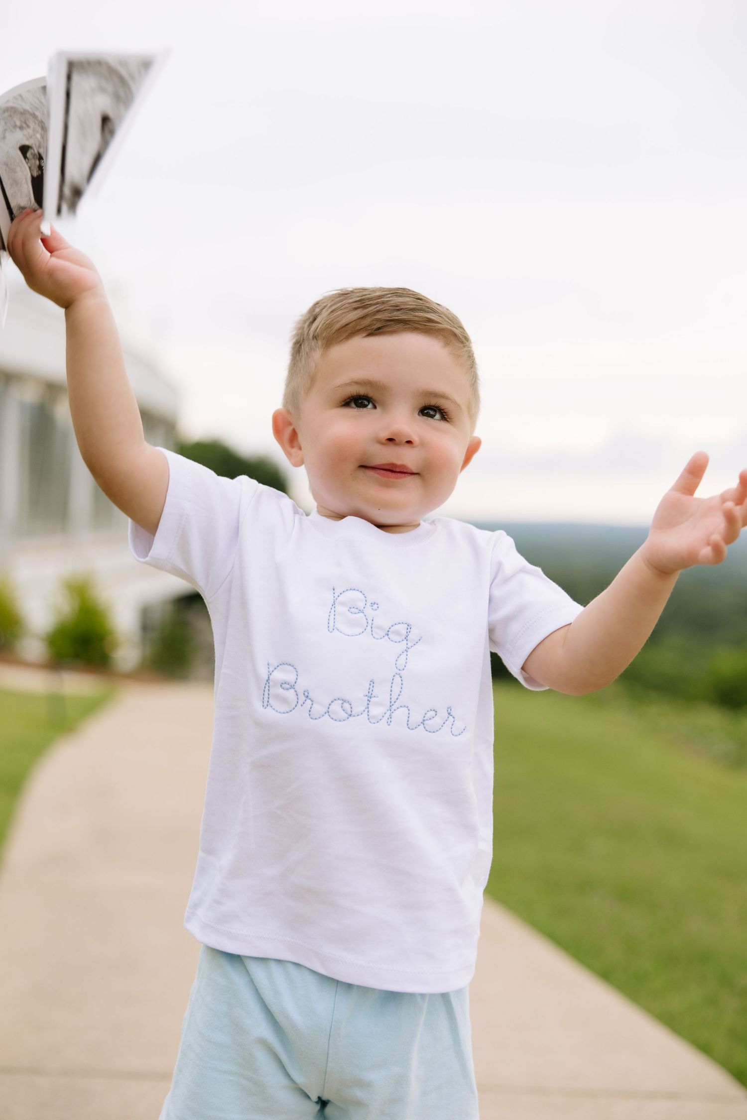 Toddler boy in white Big Brother shirt raises arms outdoors on a pathway, smiling joyfully.