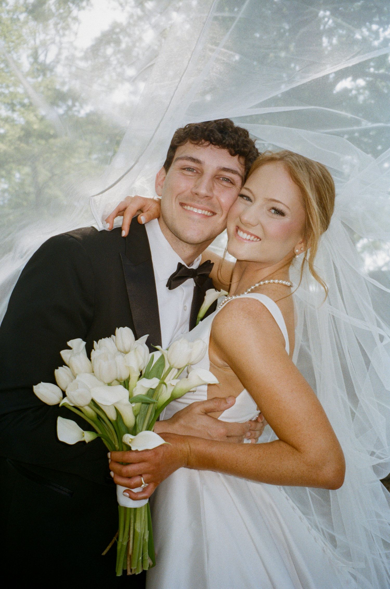 Smiling bride and groom under a flowing veil, bride holding white tulip bouquet outdoors.
