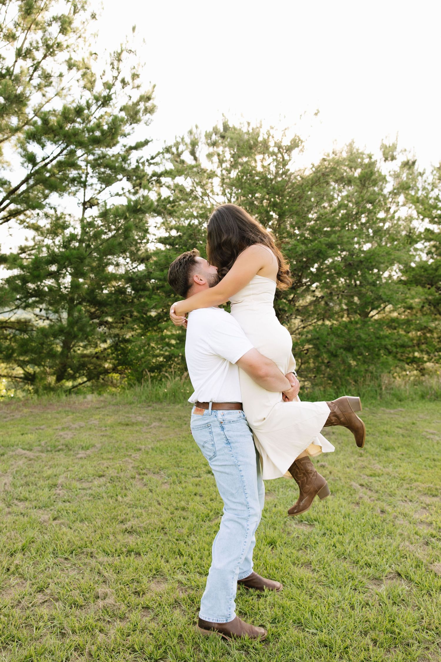 Man lifting woman in white dress and cowboy boots while kissing outdoors in a sunny green field.
