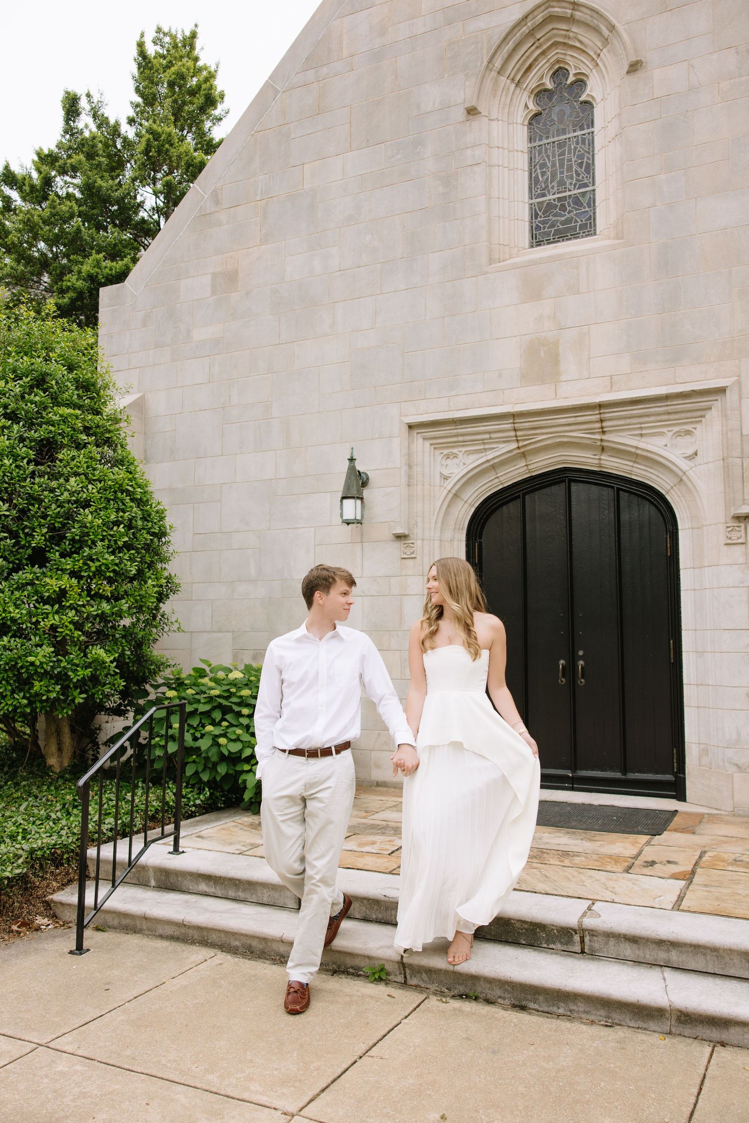 Bride and groom hold hands walking down stone church steps, bride in white strapless gown, groom in white shirt and khakis.