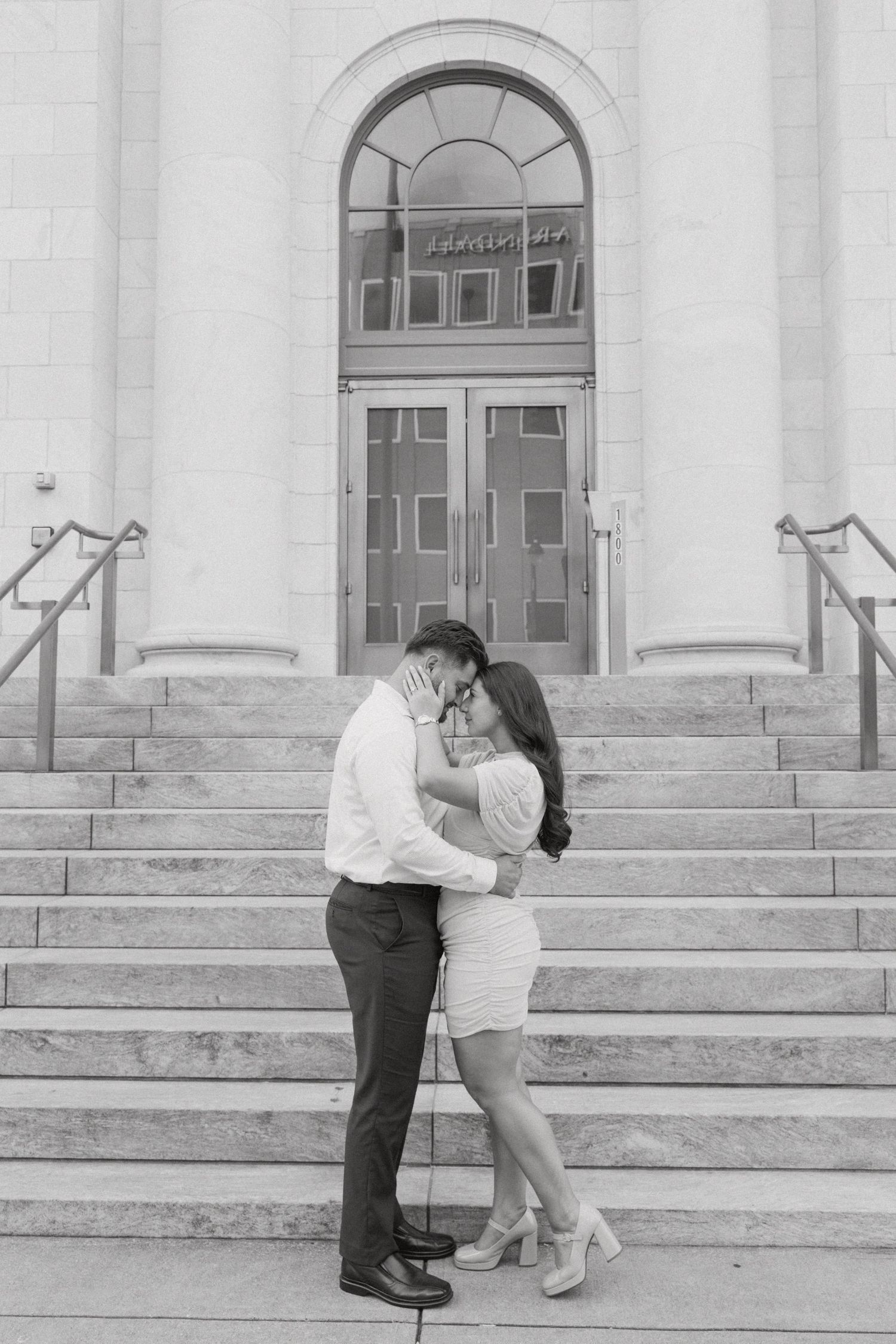 Black and white photo of a couple embracing and kissing on steps outside a grand building.