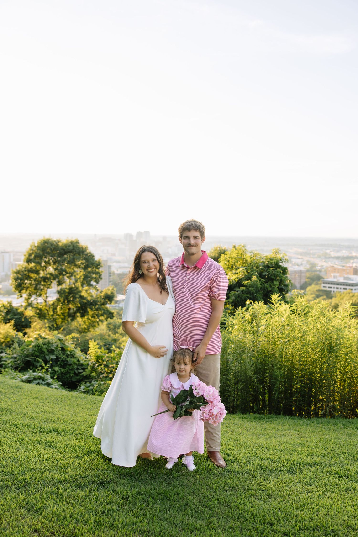 A pregnant woman in white dress poses with her husband and young daughter holding pink flowers on a green hill.