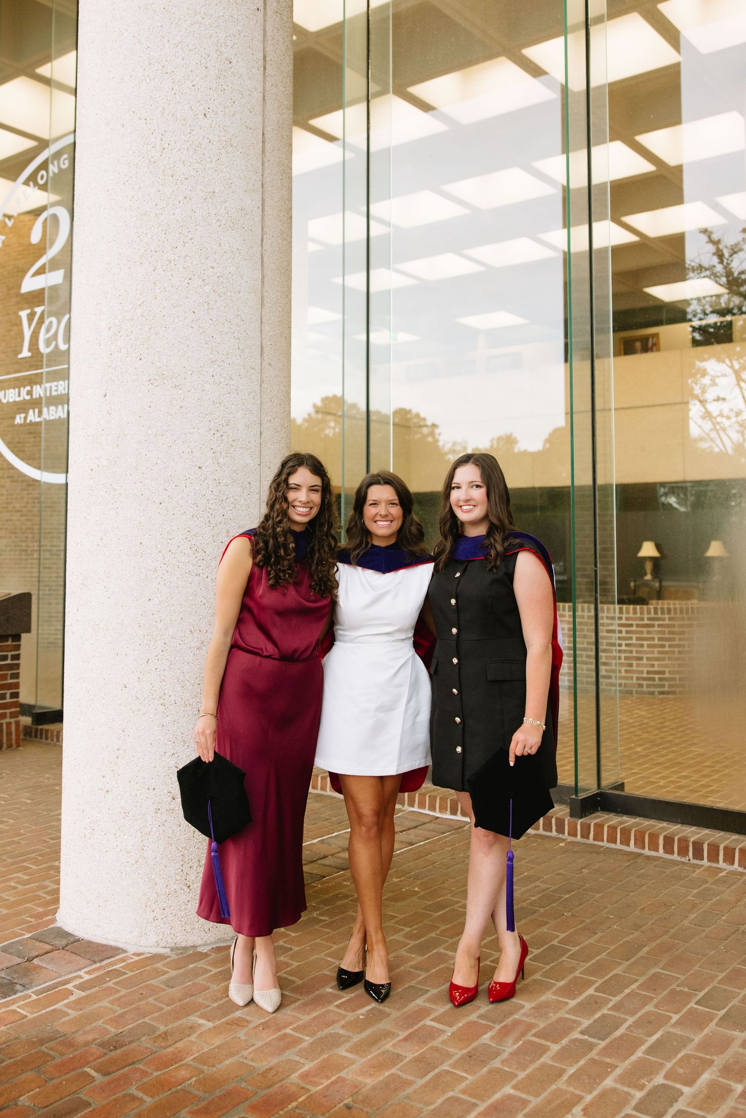 Three women in elegant dresses smile together outside a university building at a graduation celebration.