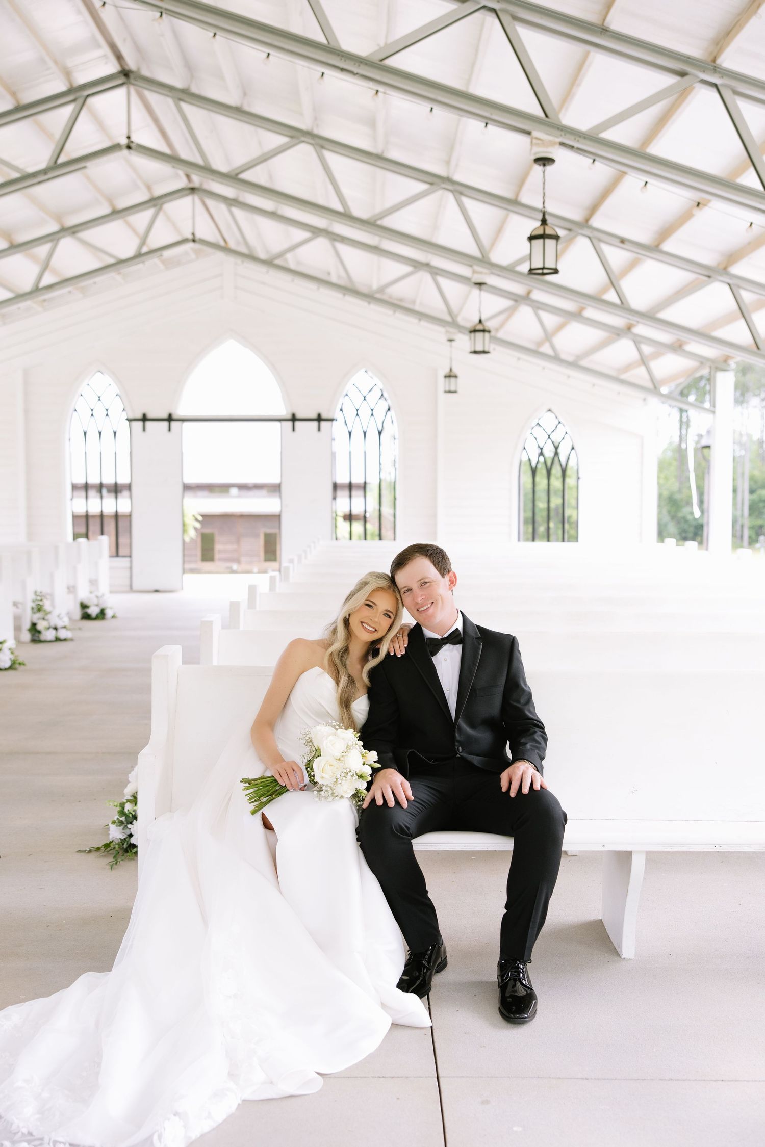 Bride and groom sitting together on white pew inside elegant white chapel with arched windows and glass ceiling.