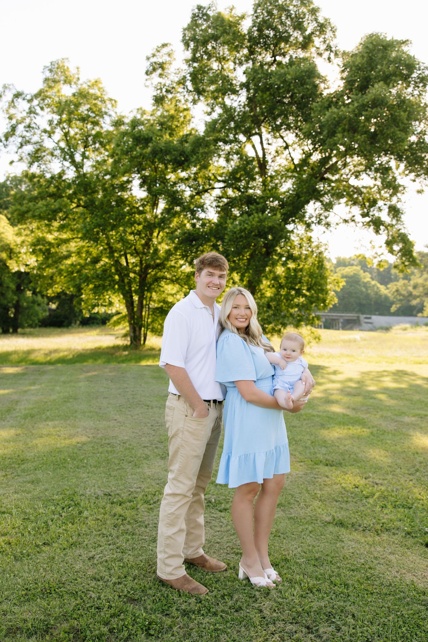 Young family of three poses outdoors on green grass, parents in light blue and white outfits holding a baby, with trees behind.