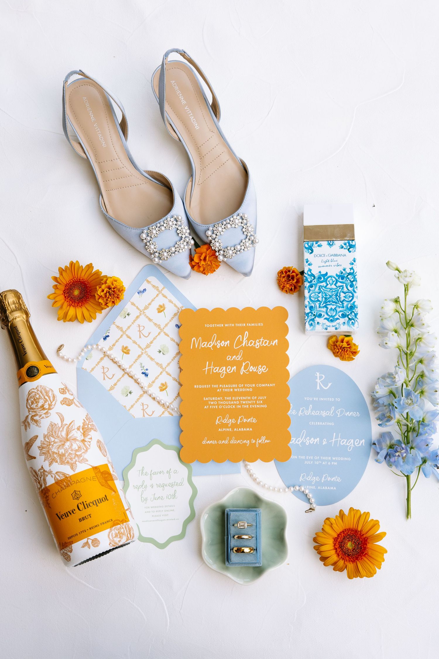 Blue heels, Veuve Clicquot champagne, orange florals, wedding invitations, rings, and stationery flatlay on white background.