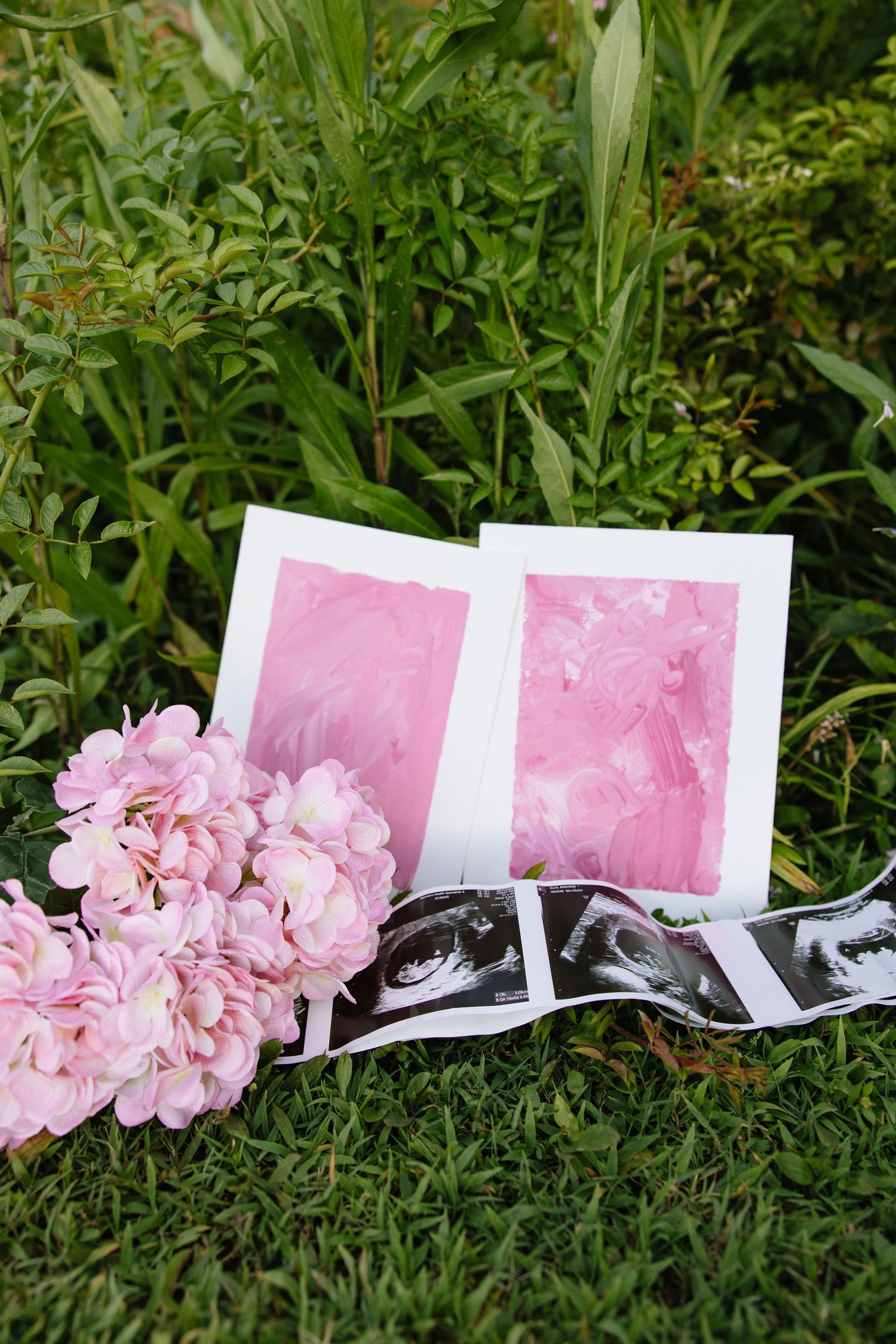 Pink watercolor art prints, ultrasound photos, and pink hydrangea flowers arranged on green grass outdoors.