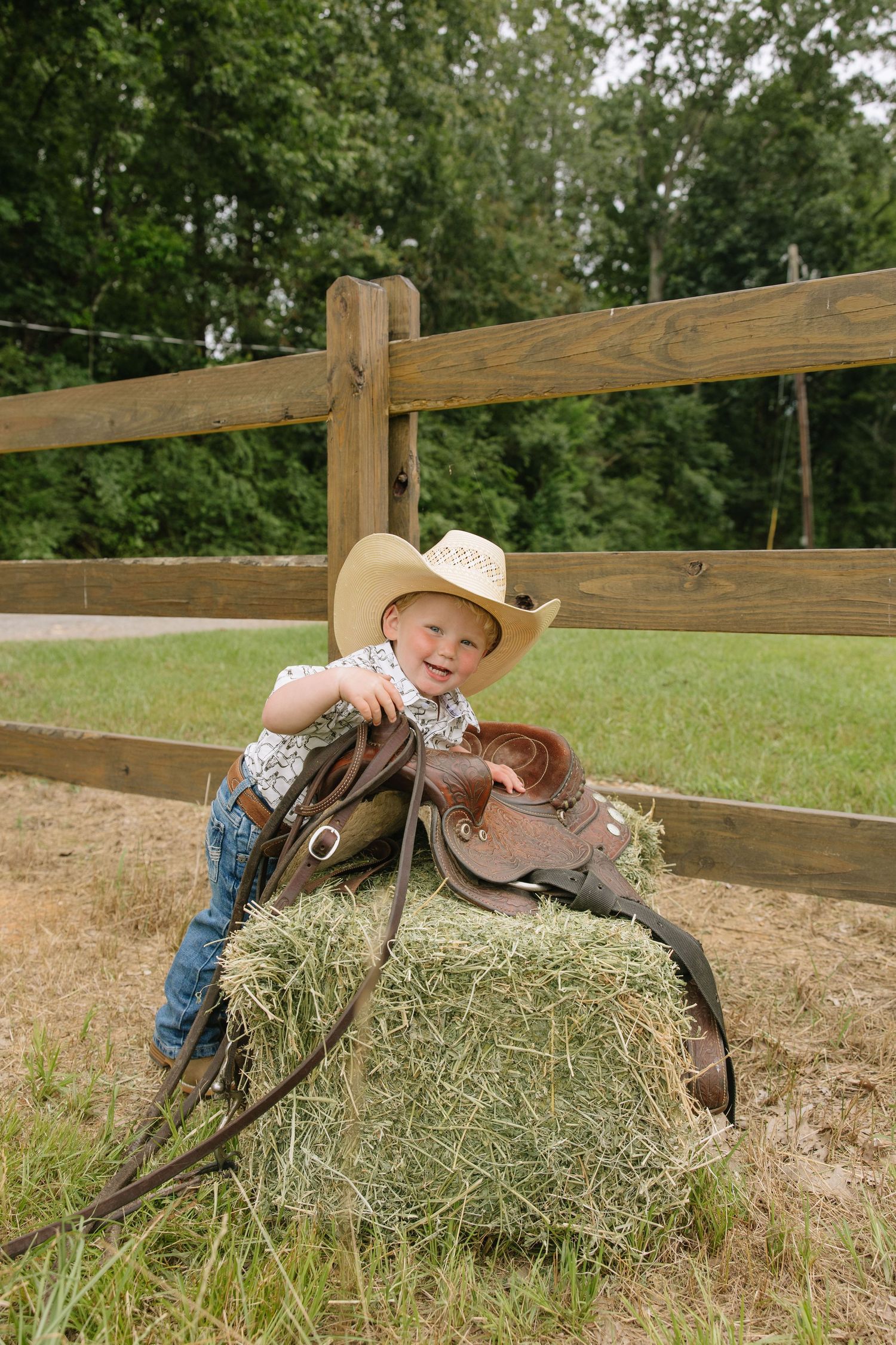 Smiling toddler in cowboy hat leans on hay bale with saddle near wooden fence outdoors.