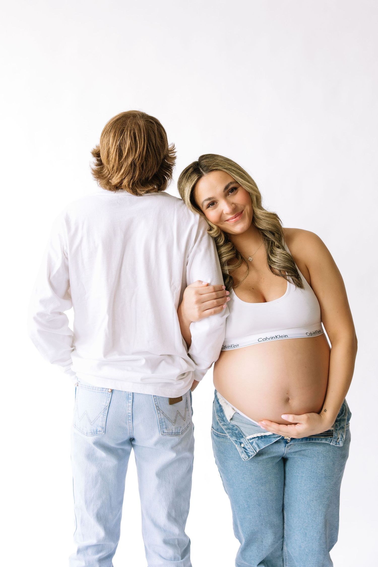 Pregnant woman in Calvin Klein sports bra and jeans poses with partner in white shirt against white background.