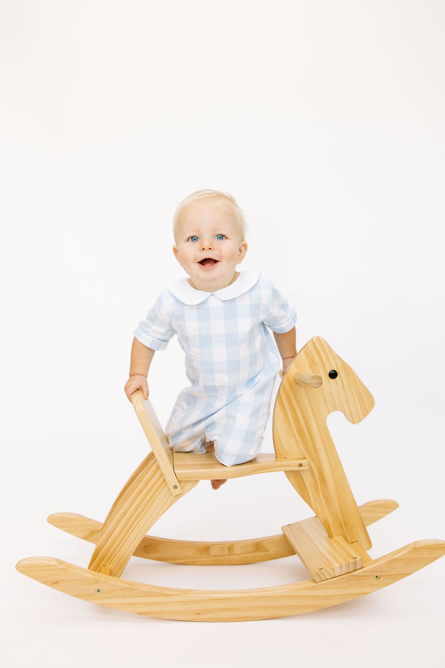 Smiling baby in blue gingham romper sitting on a wooden rocking horse against white background.