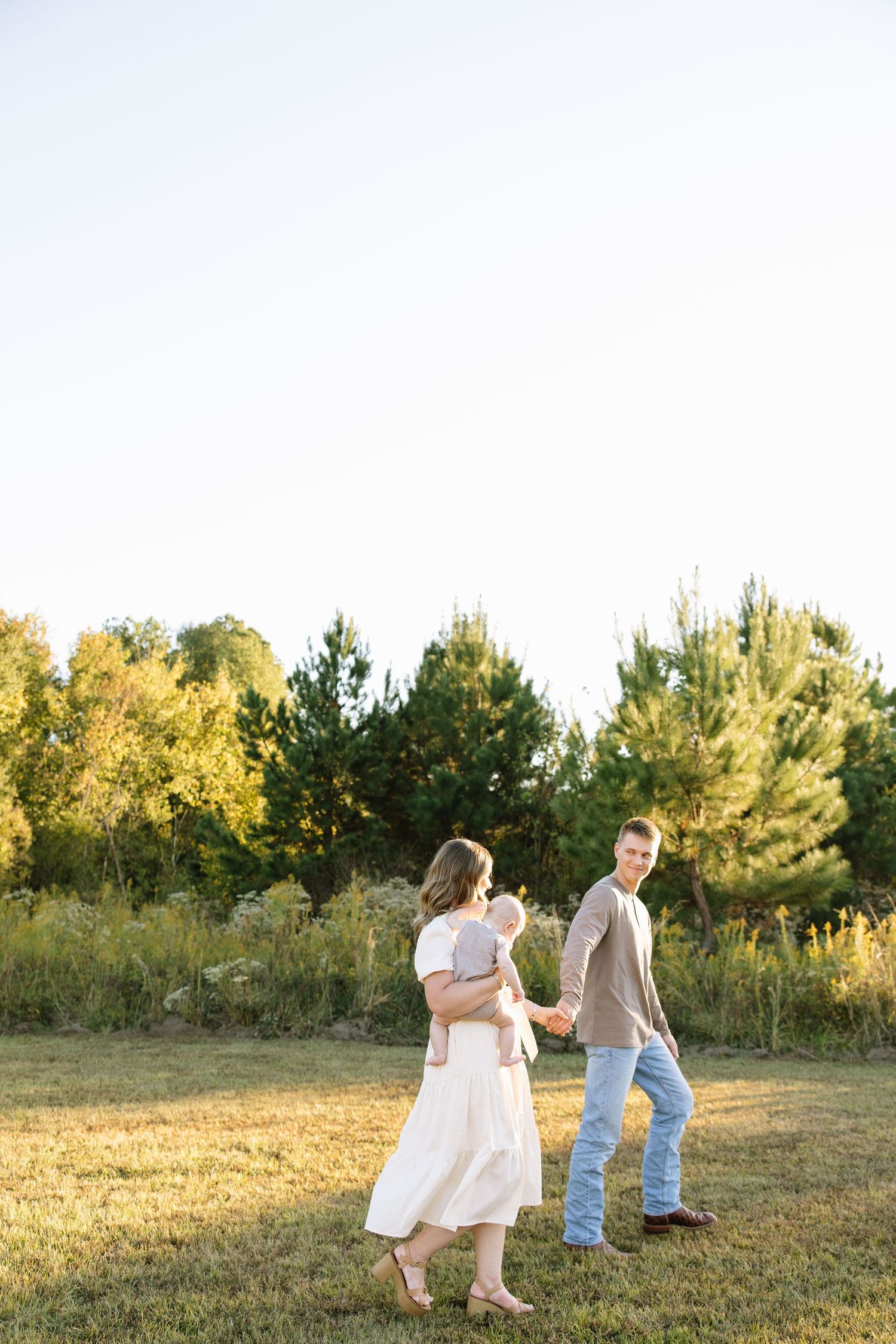 A young family walks hand-in-hand in a sunny field, with the mother holding a baby and trees in the background.
