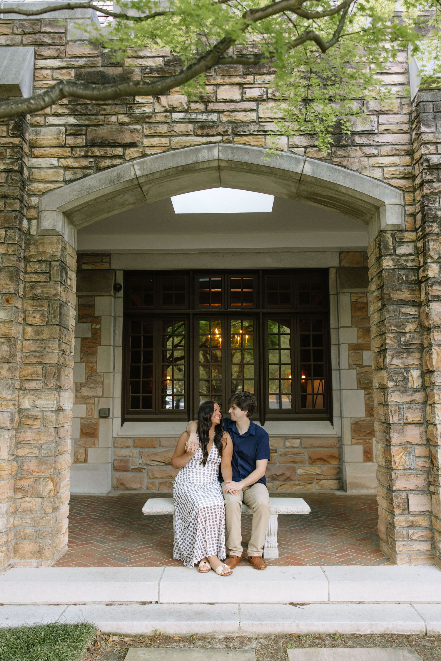 Couple sitting together on a stone bench under an arched entryway of a historic building surrounded by lush greenery.