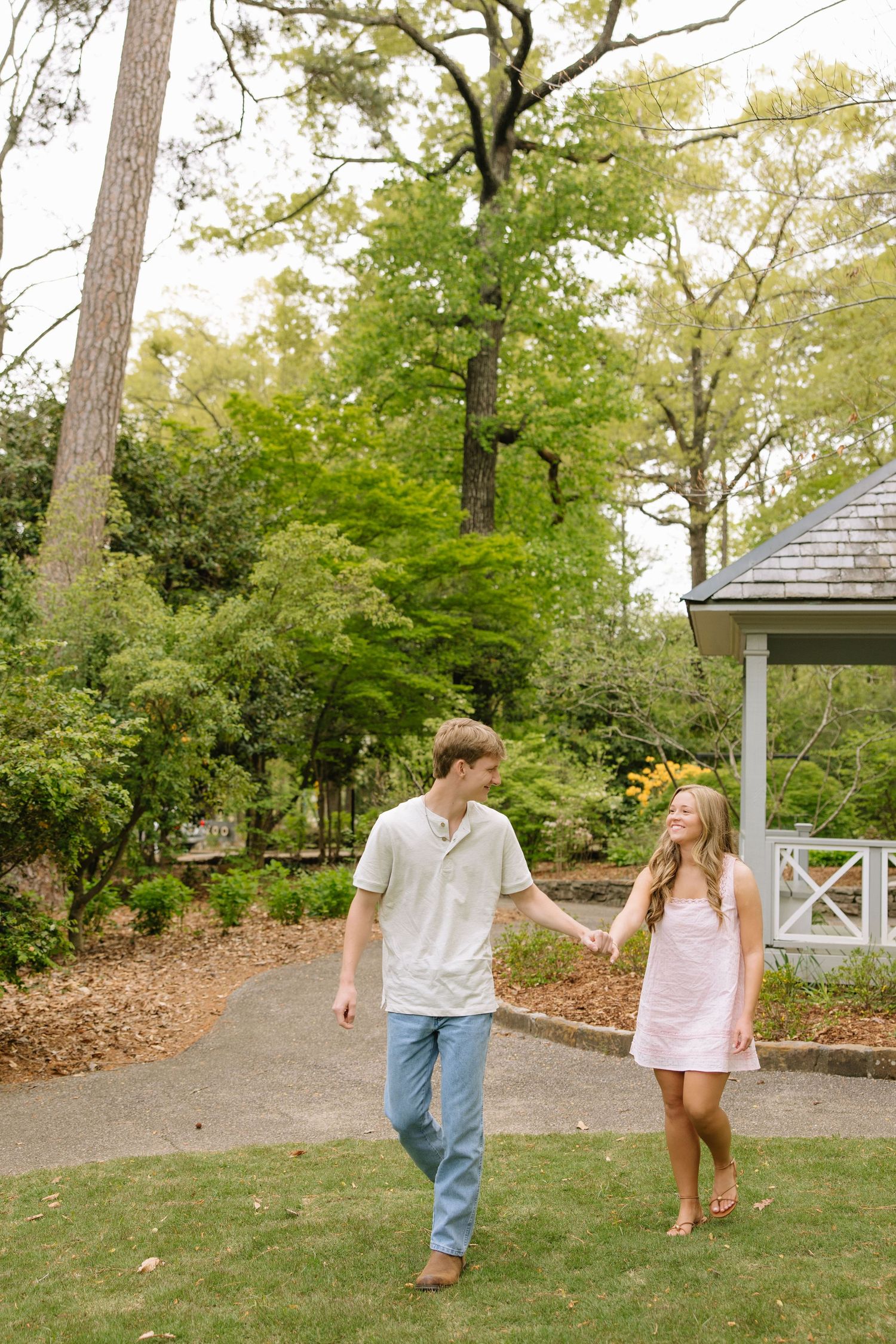 Young couple holding hands and walking together in a lush garden park near a gazebo on a sunny day.