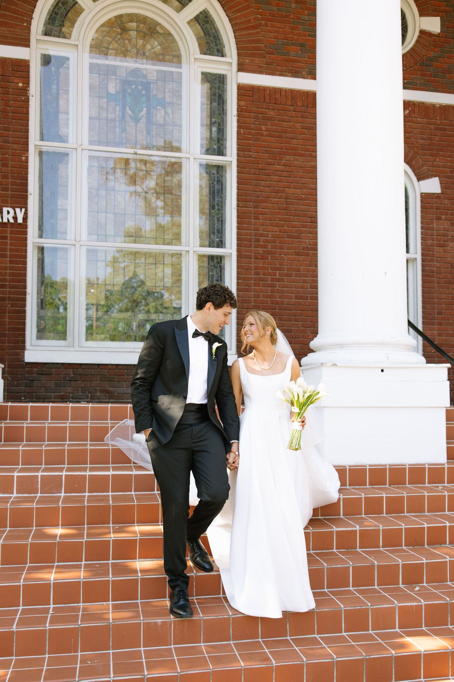 Bride and groom holding hands on red brick church steps, bride in white gown with bouquet, groom in black tuxedo.