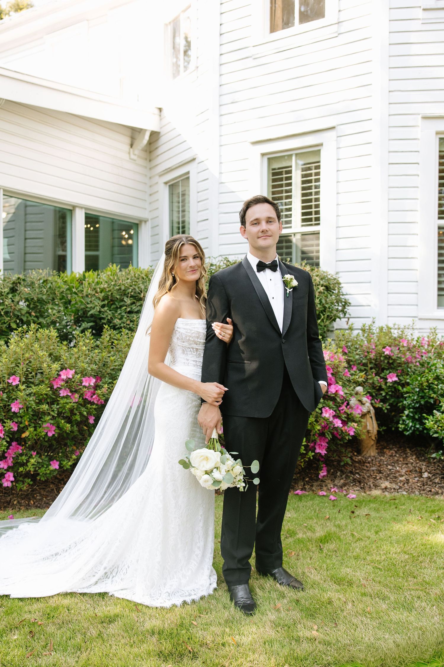 Wedding portrait in front of white house with pink flowering bushes and green shutters.