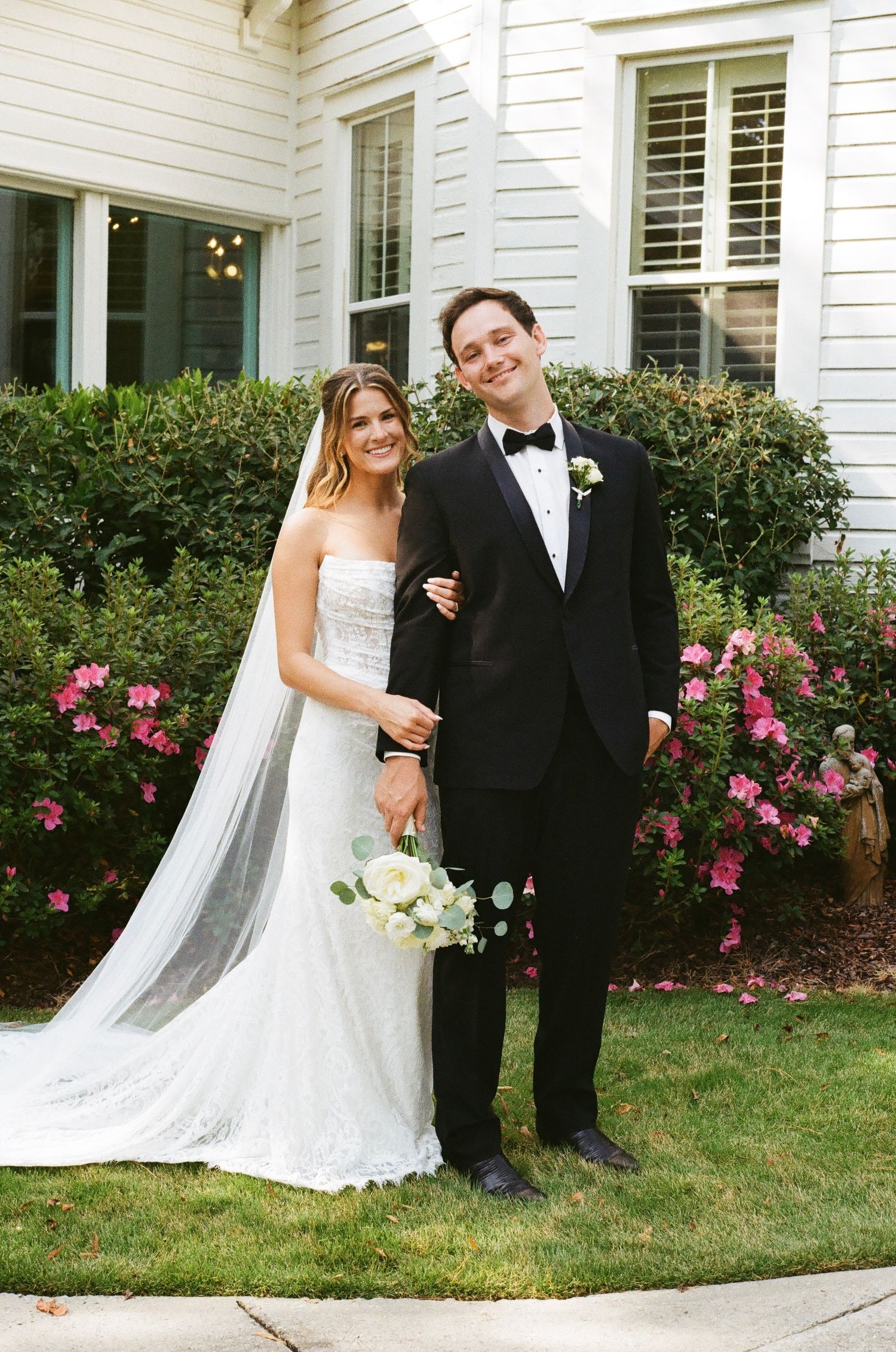 A newlywed couple poses together in front of a white house with pink rose bushes in a series of wedding photos.