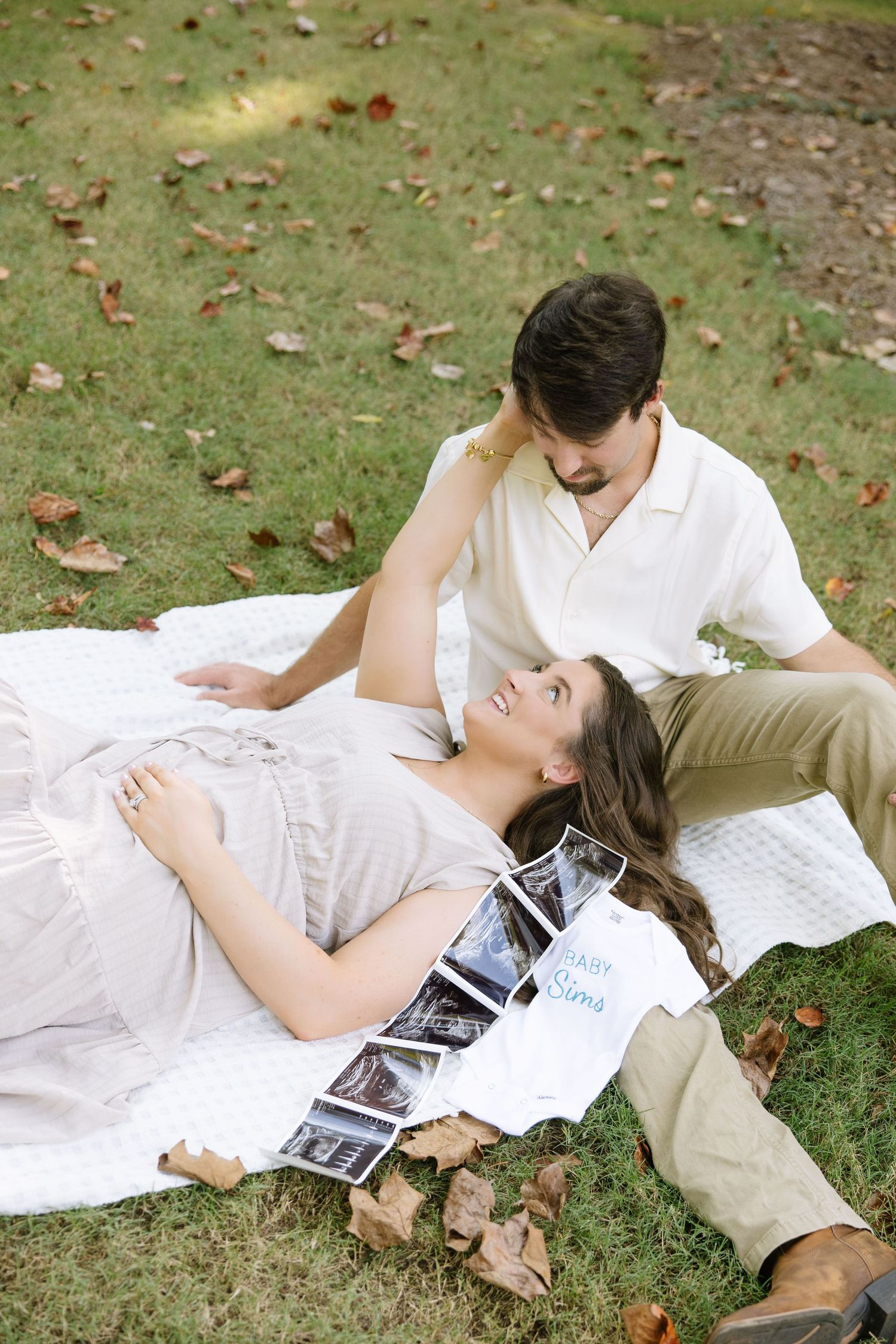 Pregnant woman lying on blanket in park with ultrasound photos, smiling up at partner sitting beside her.