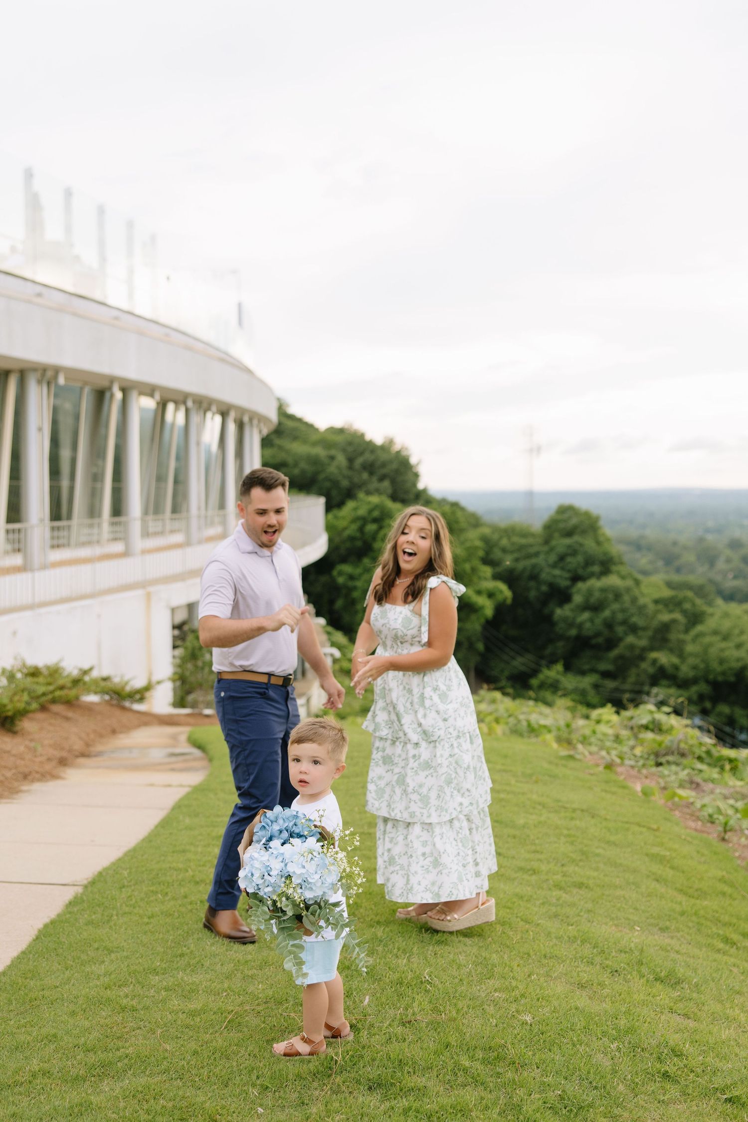 A joyful family with a toddler holding blue flowers poses on a green lawn beside an elegant white building.