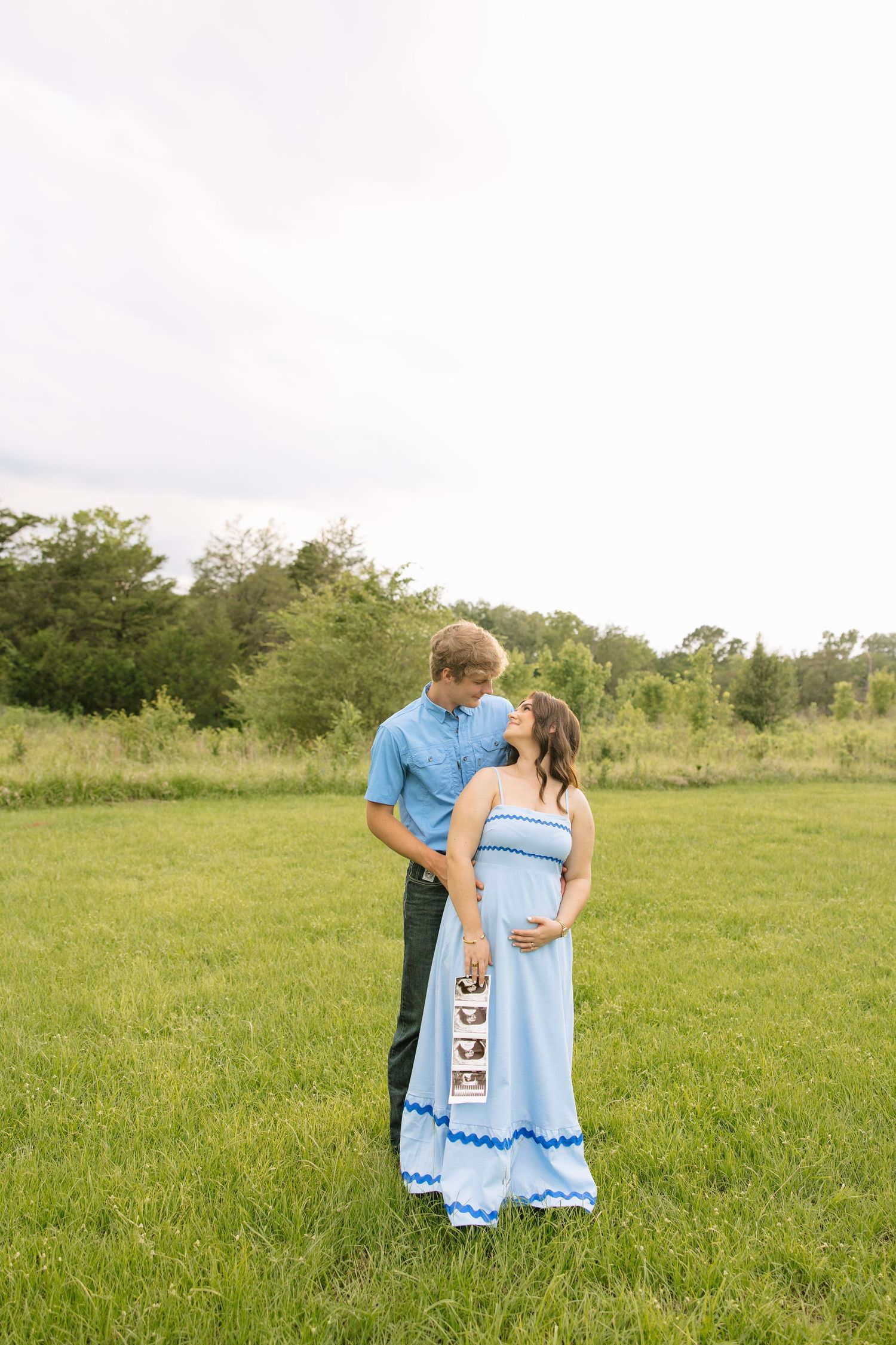 Couple in matching blue outfits standing in green field, woman in maxi dress holding ultrasound photos during maternity shoot.