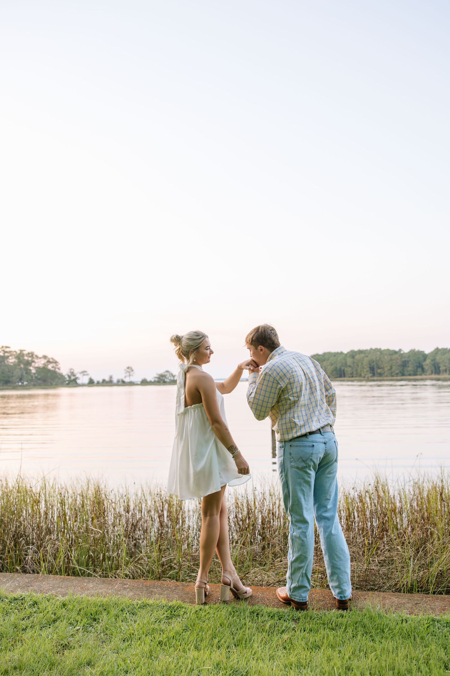 A couple shares a romantic moment by a serene lake at sunset, man kissing woman's hand near tall grass.